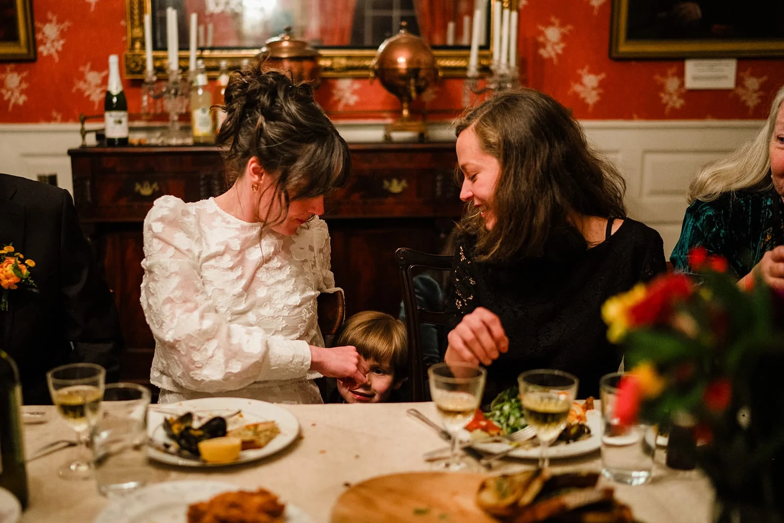A documentary photograph of a bride and her sister laughing with their nephew during the dinner portion of their Loring Greenough House Wedding.
