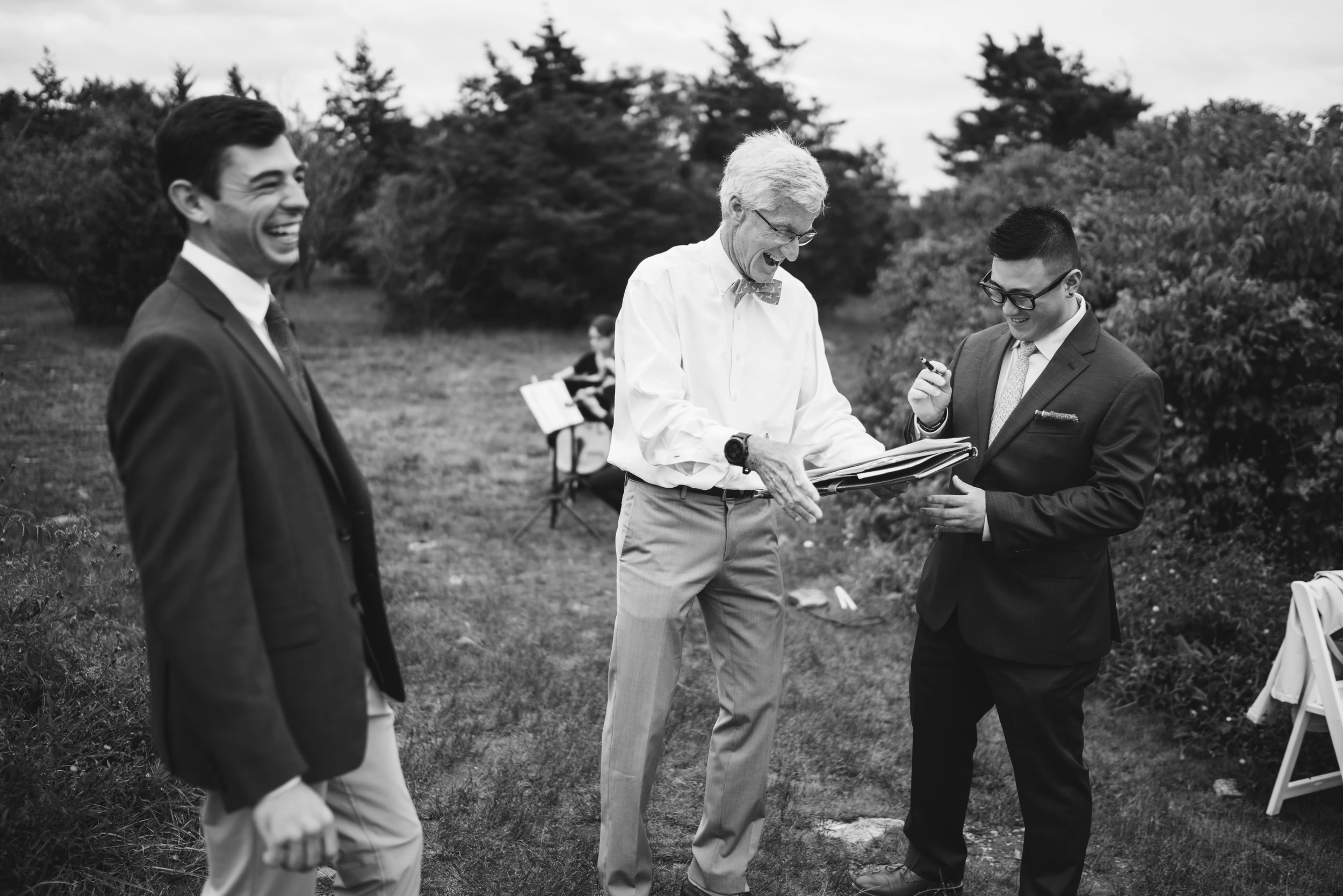 Older man in a bow tie holding an open folder, laughing with two men in suits in a grassy clearing; music stand and folding chair behind