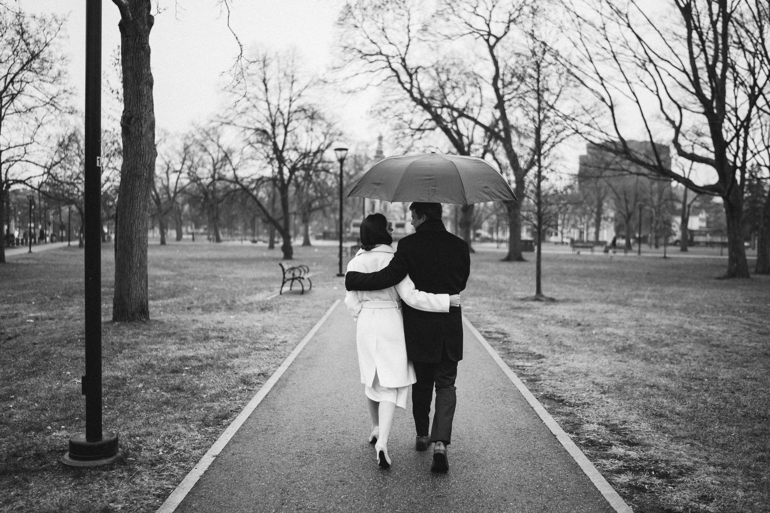 Couple walking away arm in arm under one umbrella down a straight park path, bare winter trees on both sides