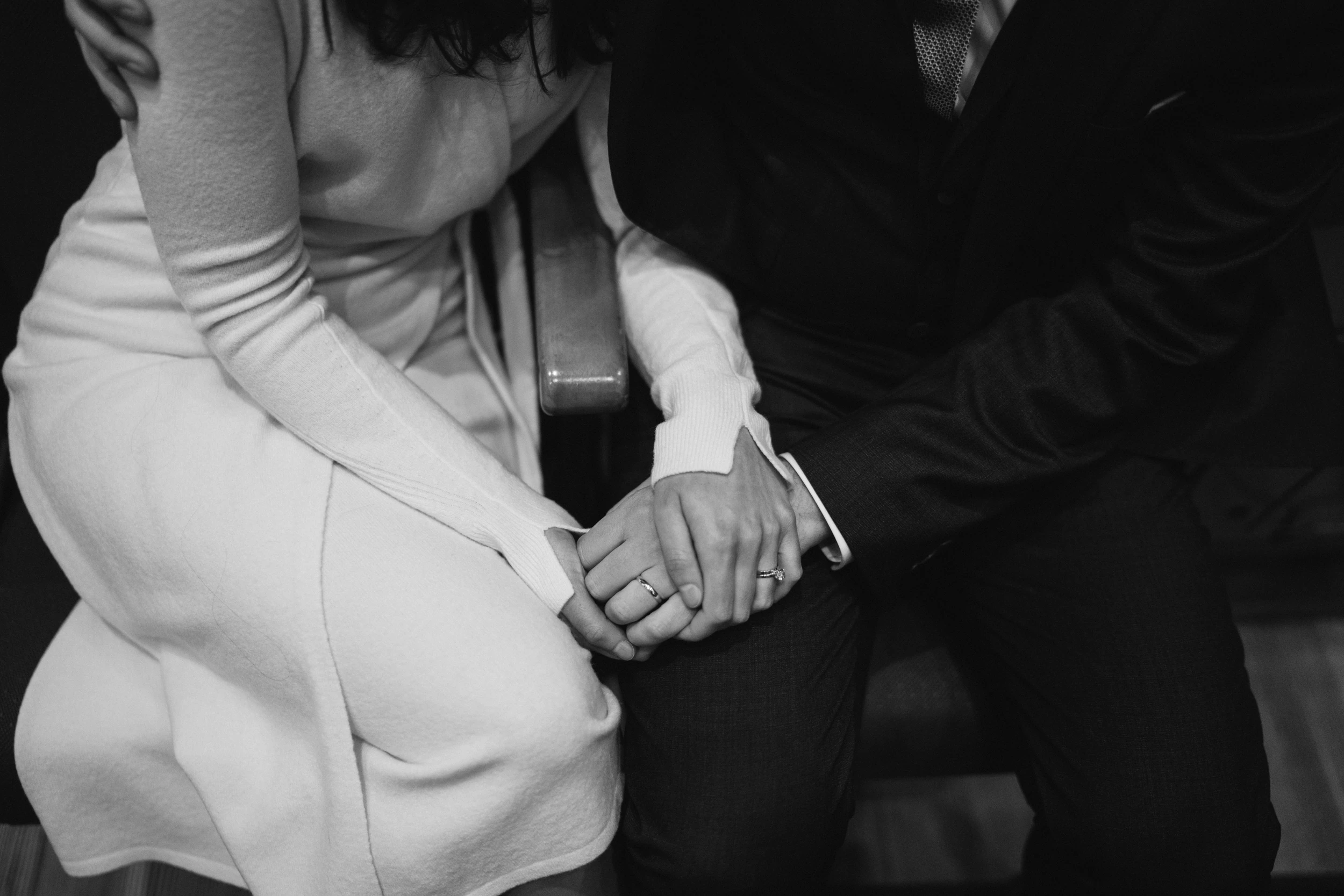 Close-up of a couple's joined hands with rings, white coat sleeve against a dark suit