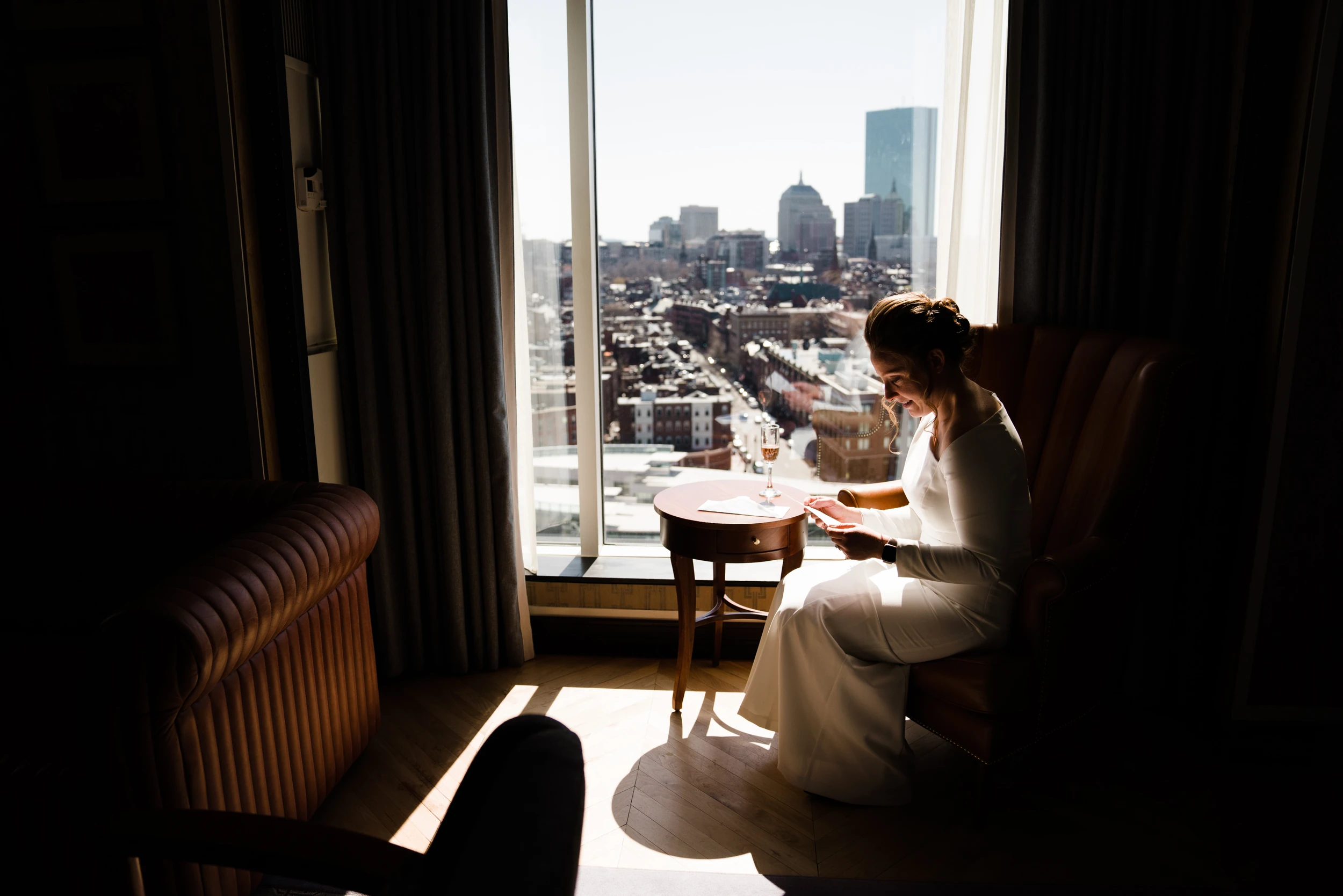 Bride in white reading a card in an armchair beside a tall window, city skyline beyond, room in deep shadow