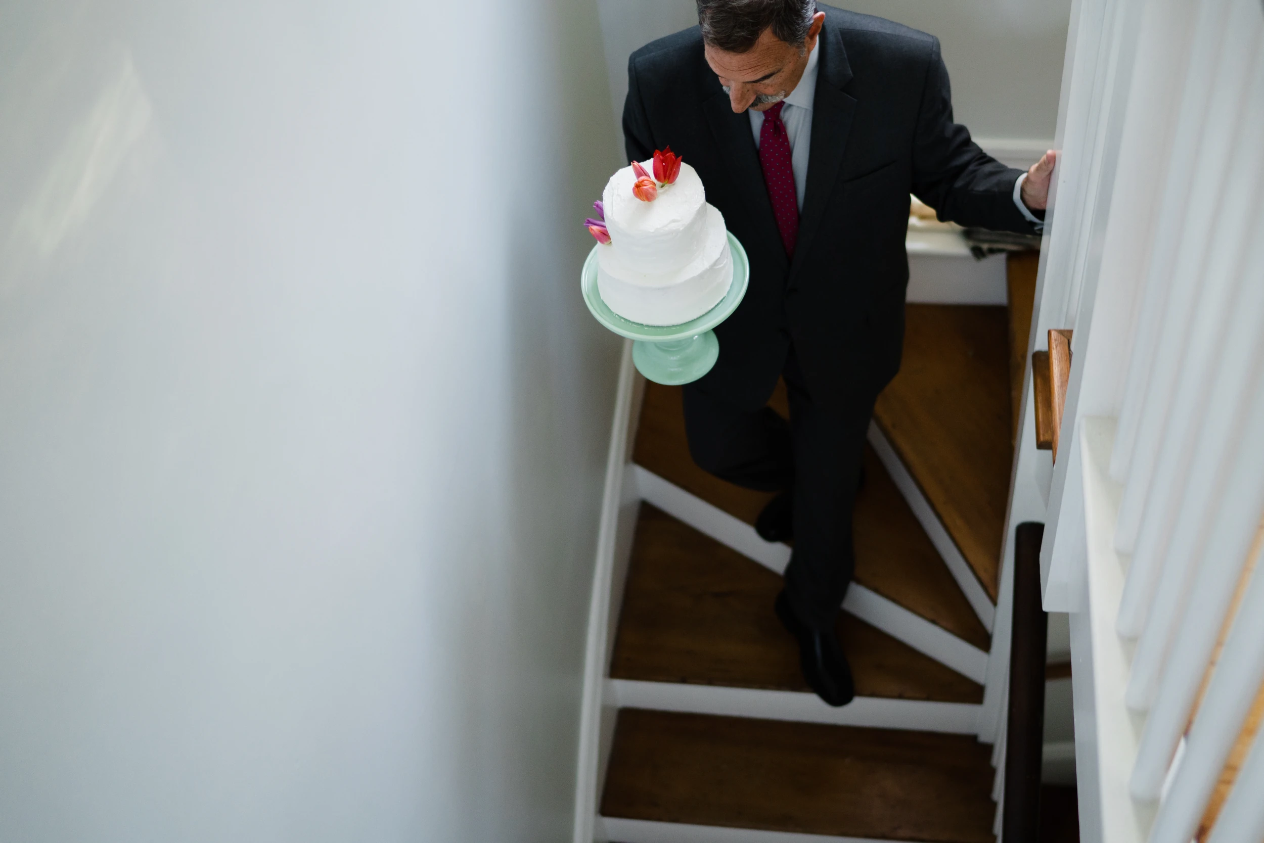 Overhead view of a man in a suit carrying a white tiered cake down a narrow staircase, bare pale wall filling the left half
