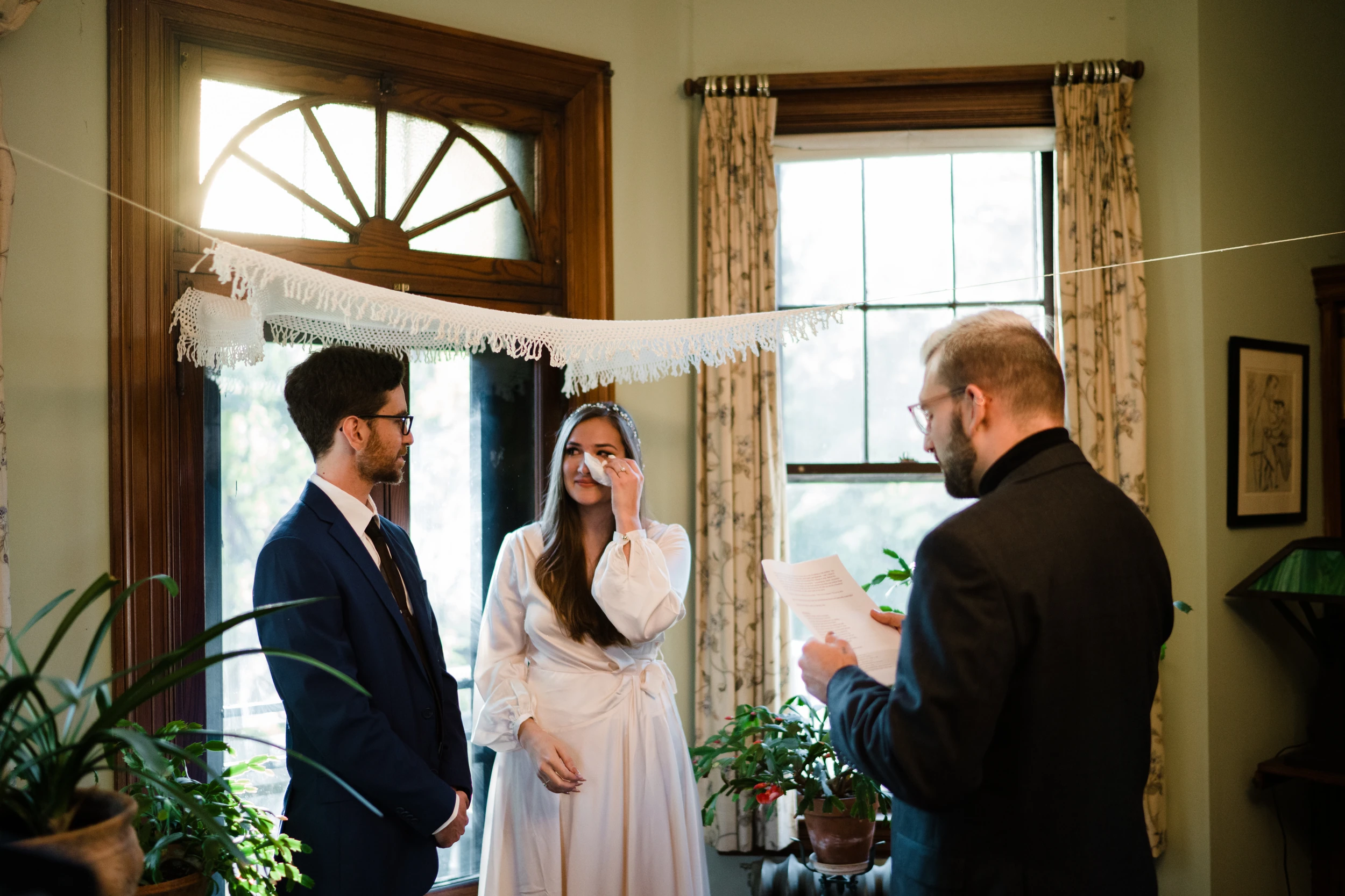 Bride wipes a tear beside the groom while a man reads from a page, under a fringed cloth strung across a living-room window