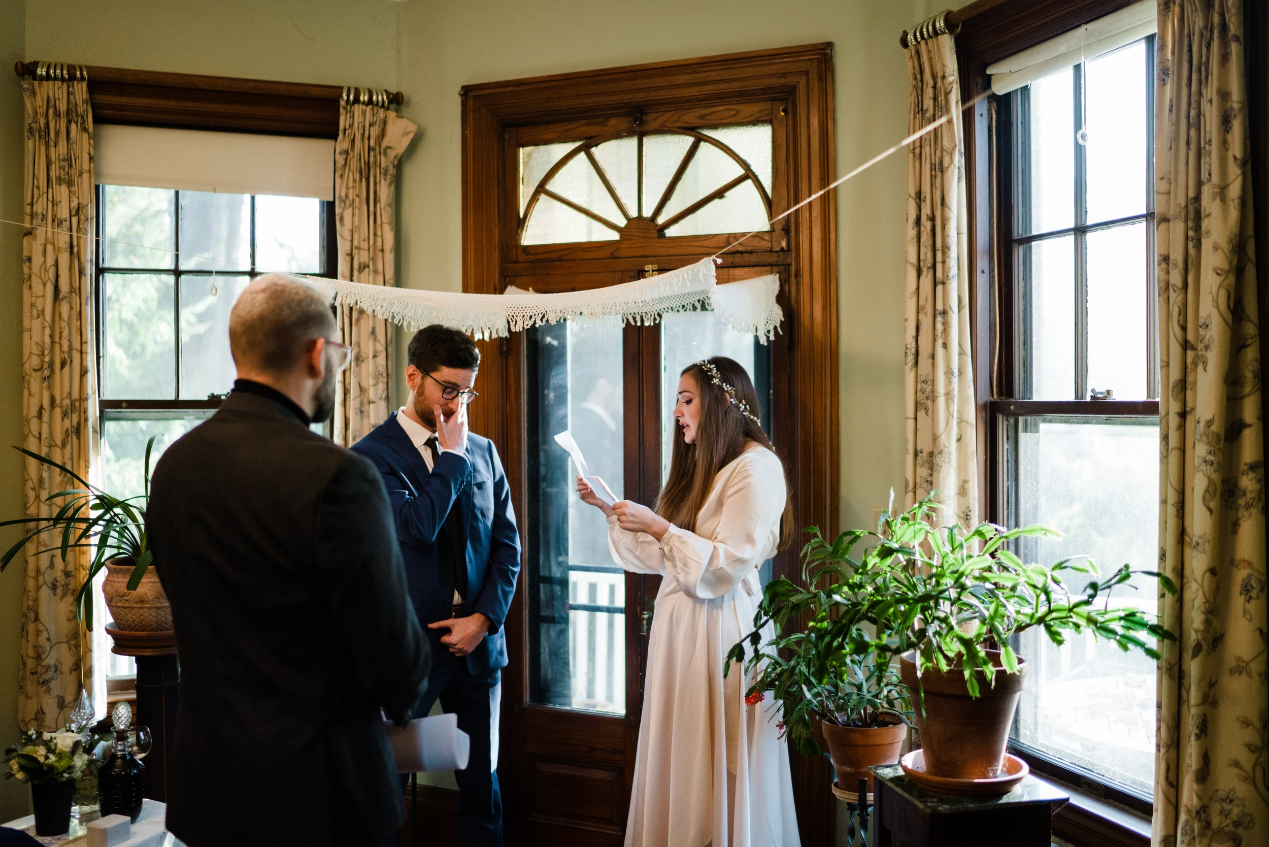 Bride reads from a card while the groom covers his mouth, a man seen from behind in the foreground, same living-room ceremony