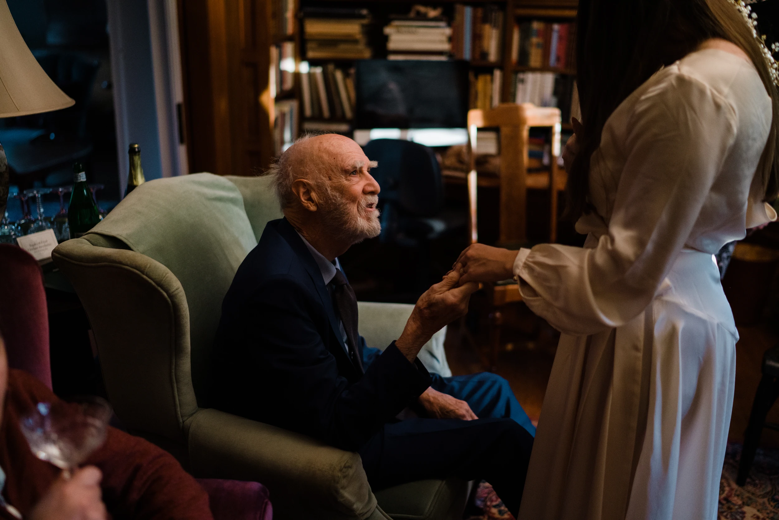 Elderly man in a green armchair holding the bride's hand, bookshelves and lamplight behind