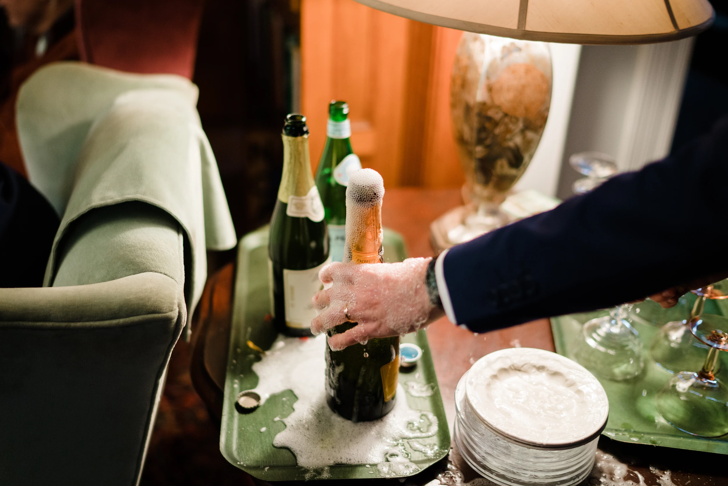 Hand gripping an overflowing champagne bottle on a green tray beside a lamp and stacked plates