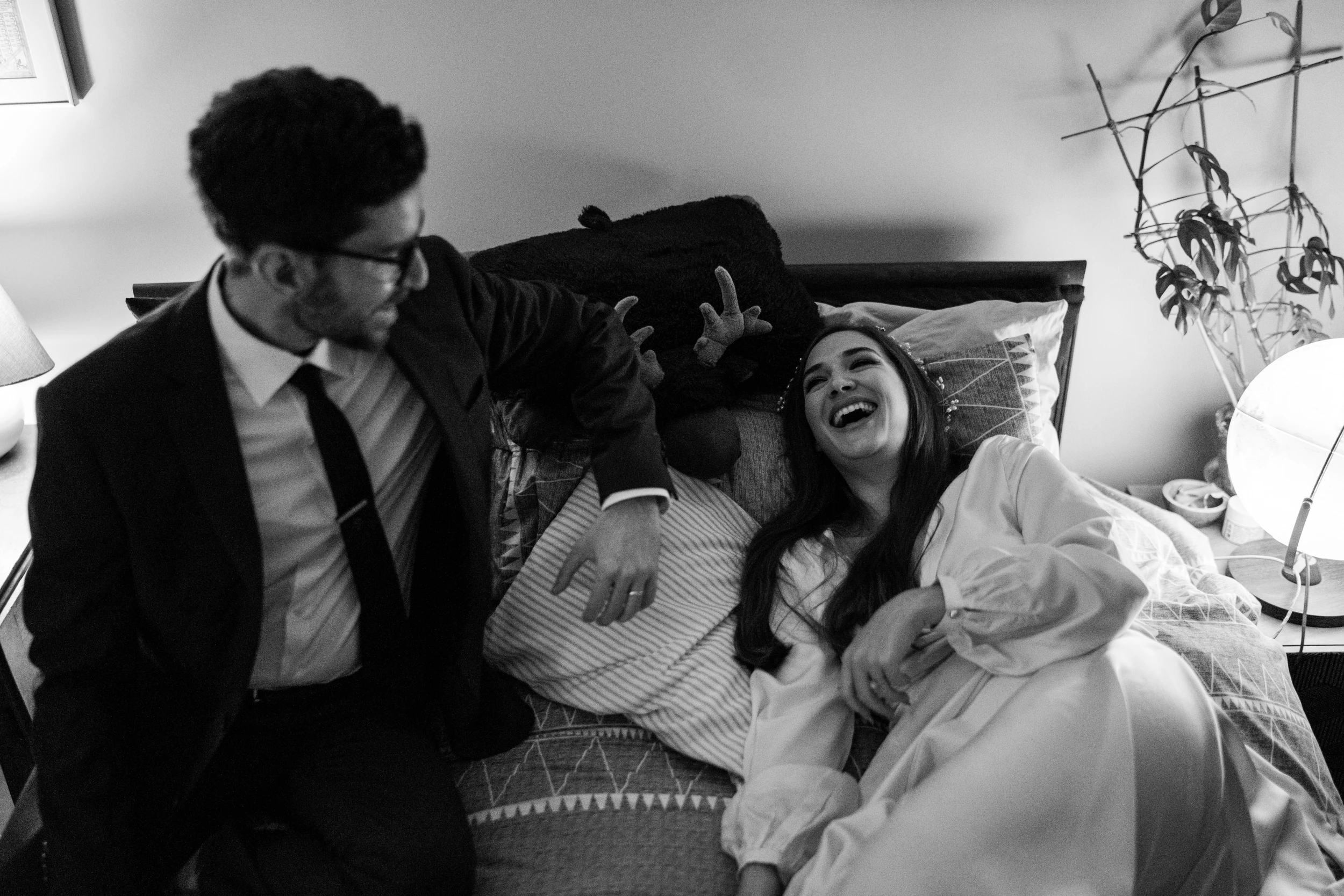 Bride laughing as she lies back on a bed, groom in a suit sitting beside her