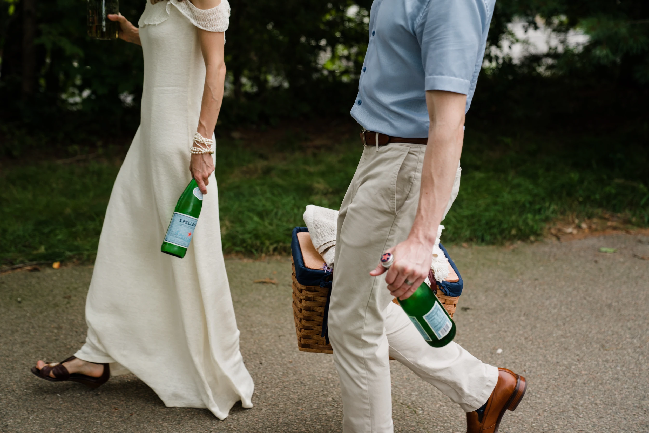 Couple cropped at the shoulders walking on a park path, carrying glass water bottles and a picnic basket