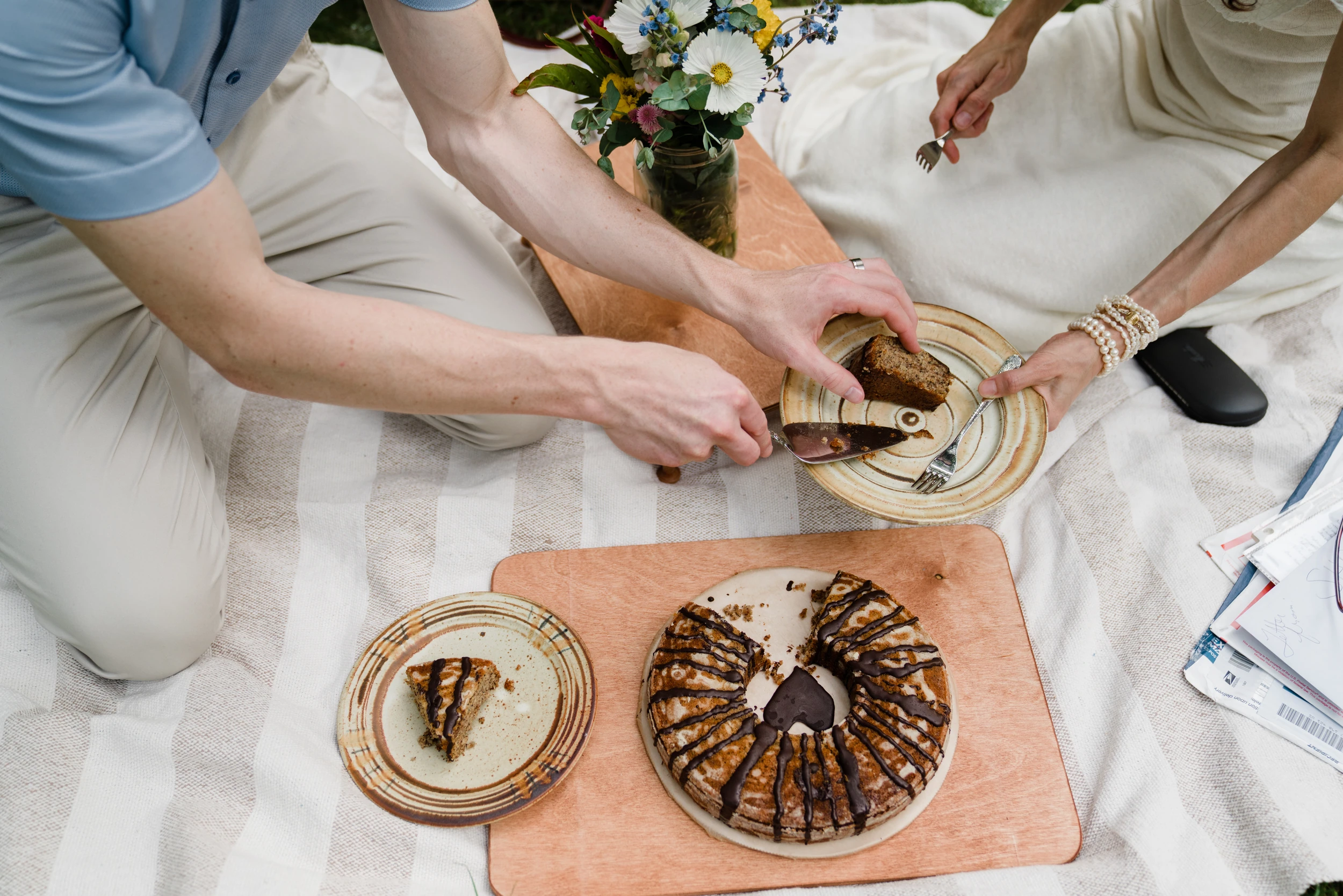 Overhead of hands serving slices of a chocolate-drizzled bundt cake on a striped picnic blanket, wildflowers in a jar