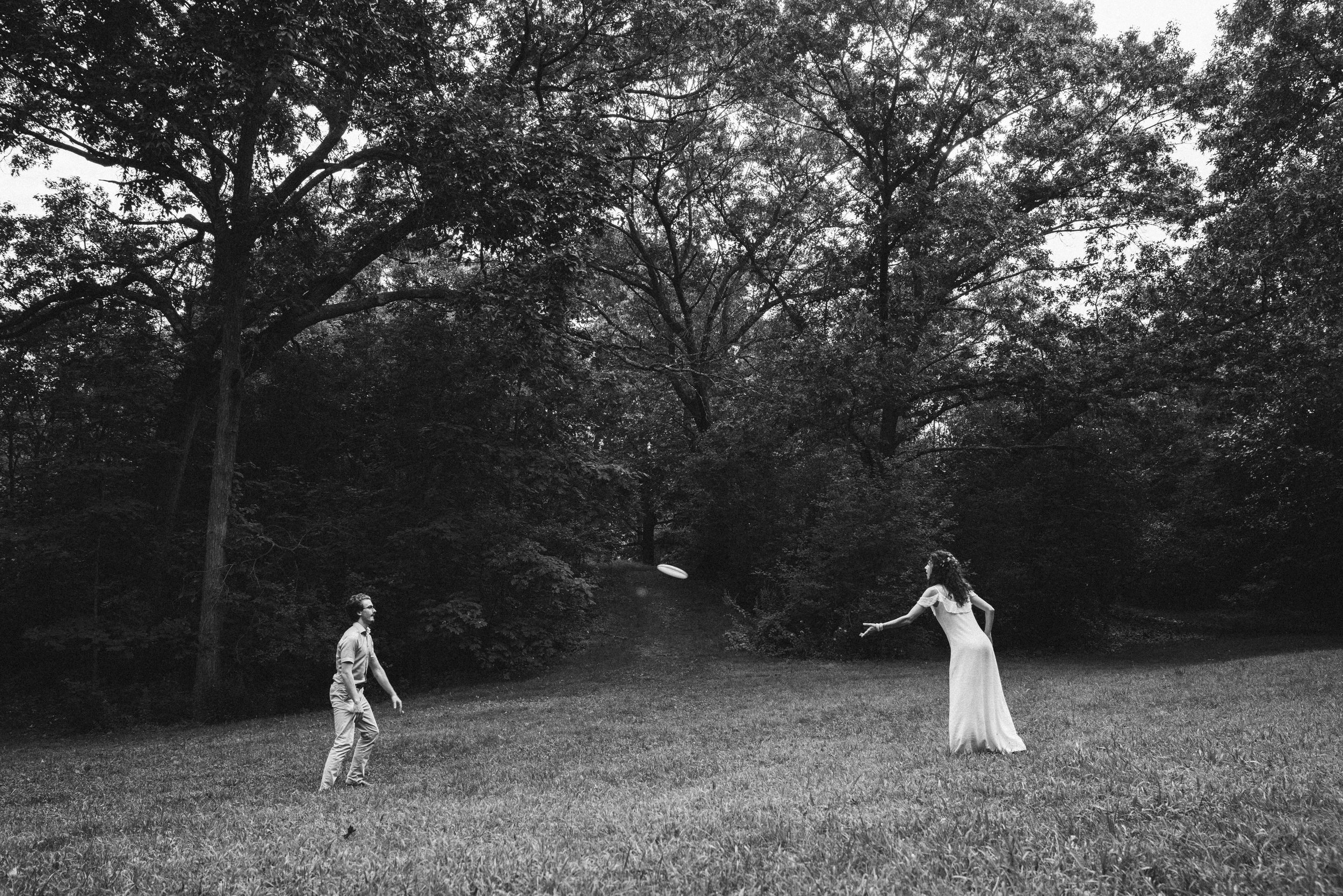 The couple far apart on a park lawn, playing catch under tall trees