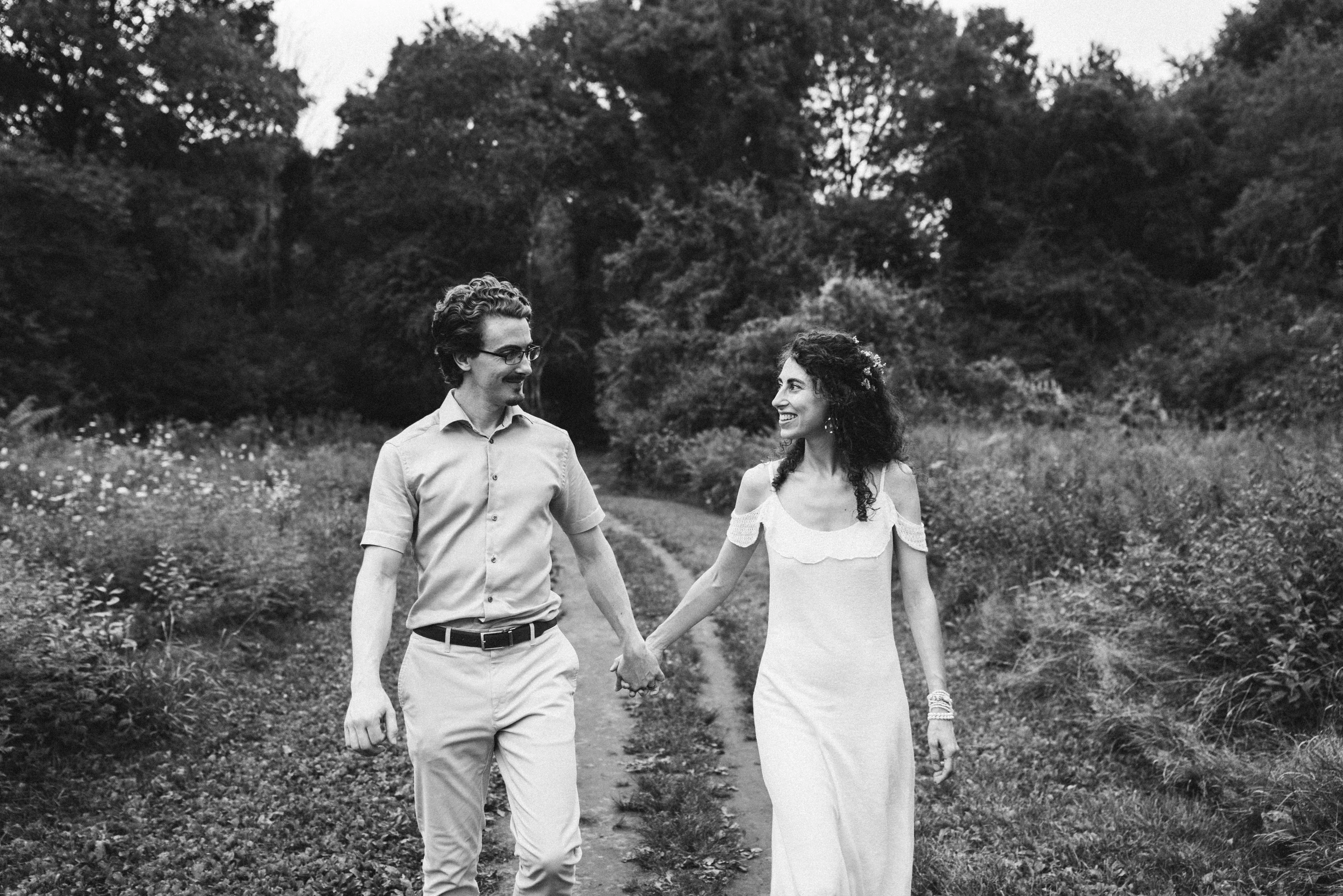 Couple walking hand in hand toward the camera on a dirt track through a meadow, smiling at each other