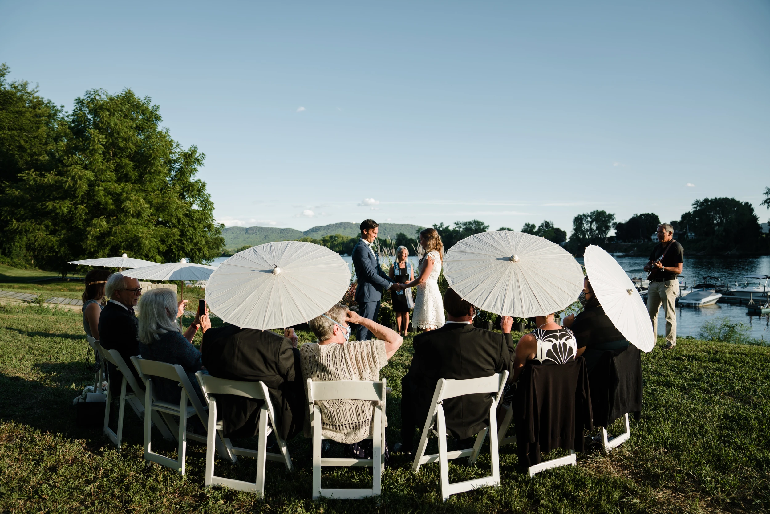 Guests on white chairs under paper parasols by the water