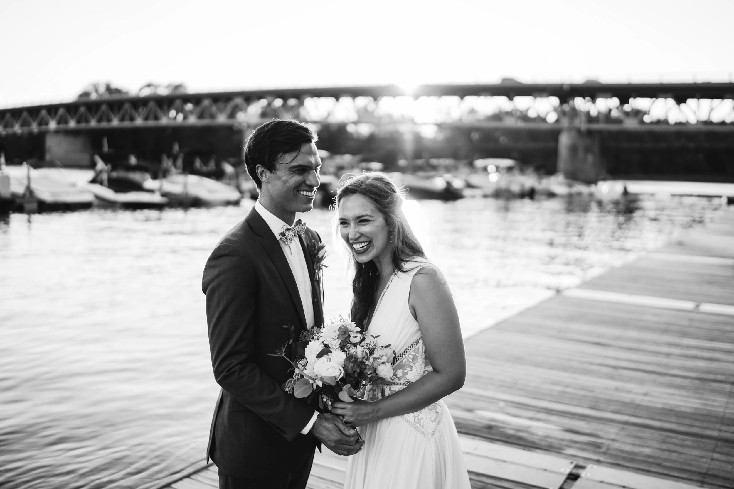 The couple by the water with a bridge behind them