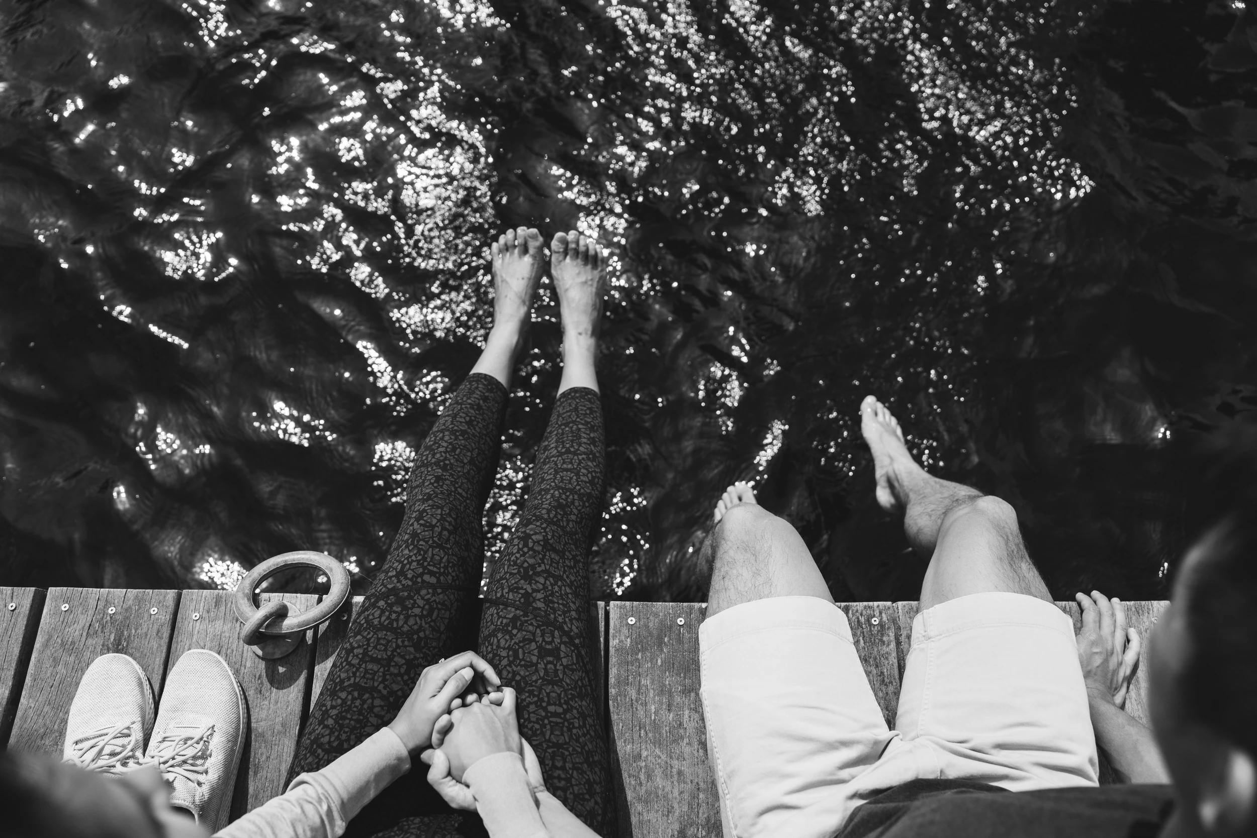 Overhead of a couple's legs and bare feet over the edge of a dock above sparkling dark water, hands clasped
