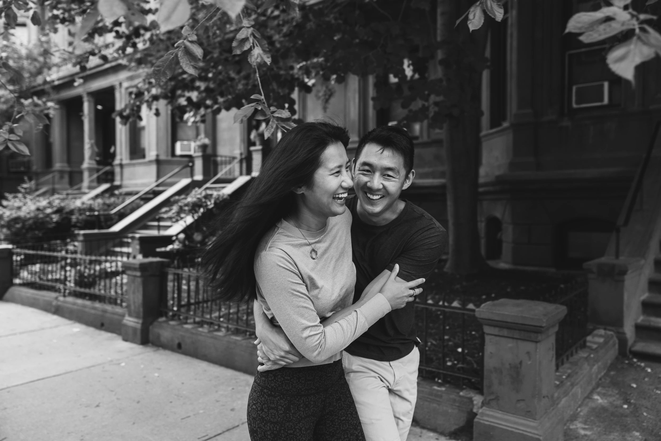 Couple laughing and embracing on a sidewalk in front of brownstone stoops, her hair blowing