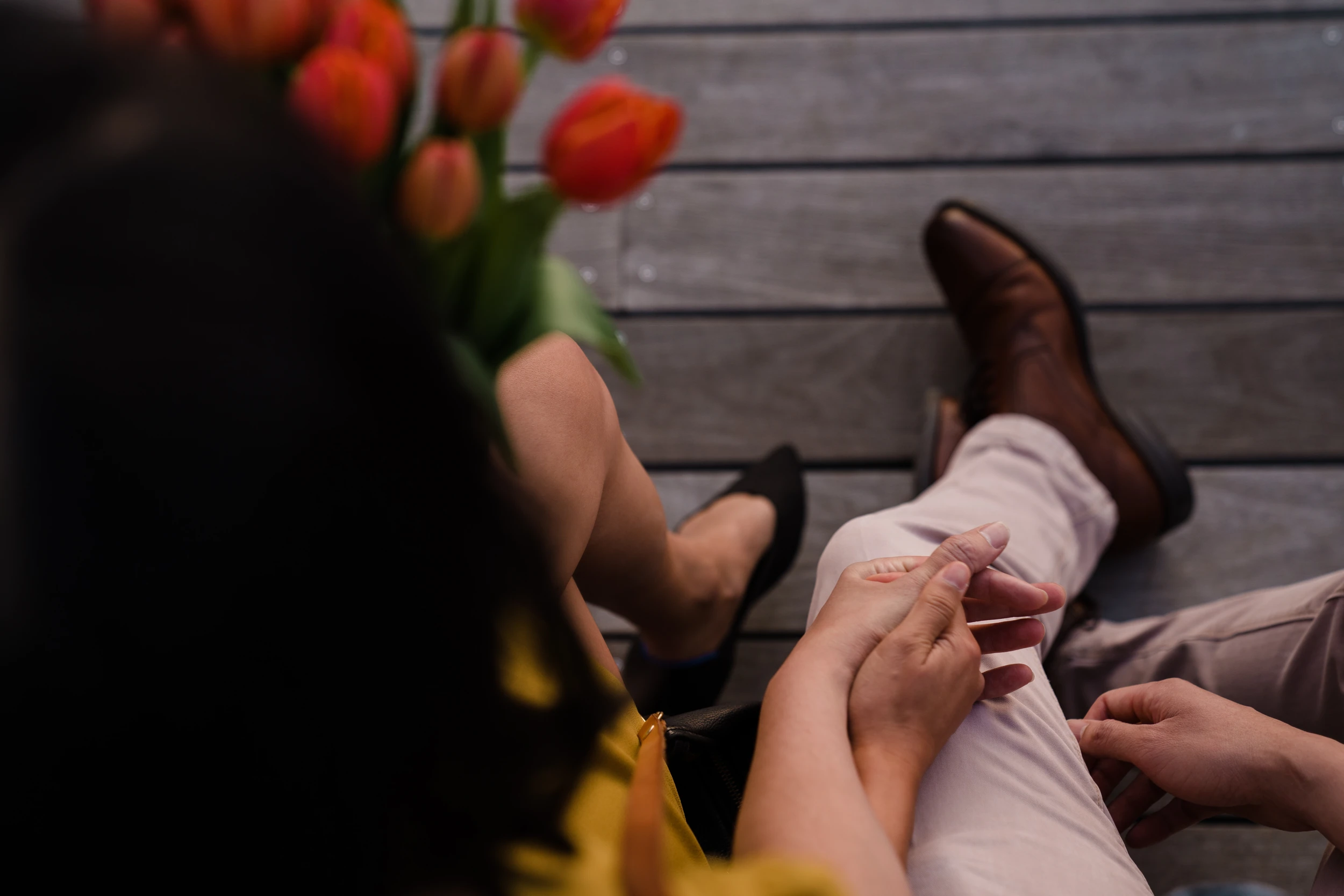 From above, a couple's hands touching over crossed legs on a wooden deck, orange tulips in the corner