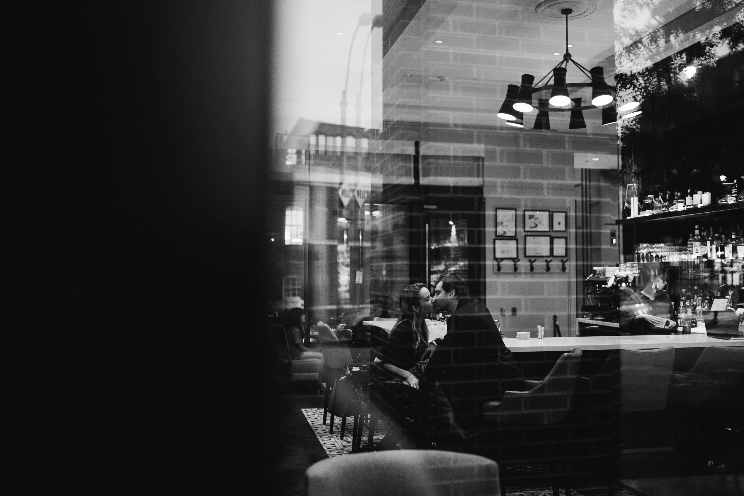 Couple kissing at a restaurant bar seen through a reflective window, left third of the frame black