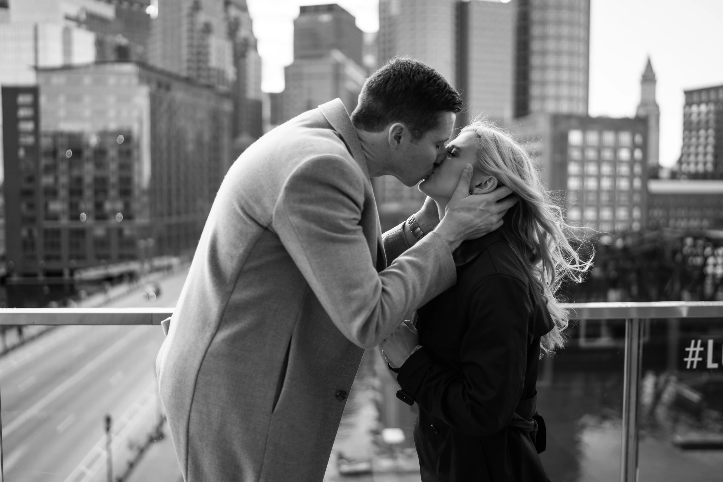 Man holds the woman's face as they kiss at a balcony railing, city buildings and a highway behind