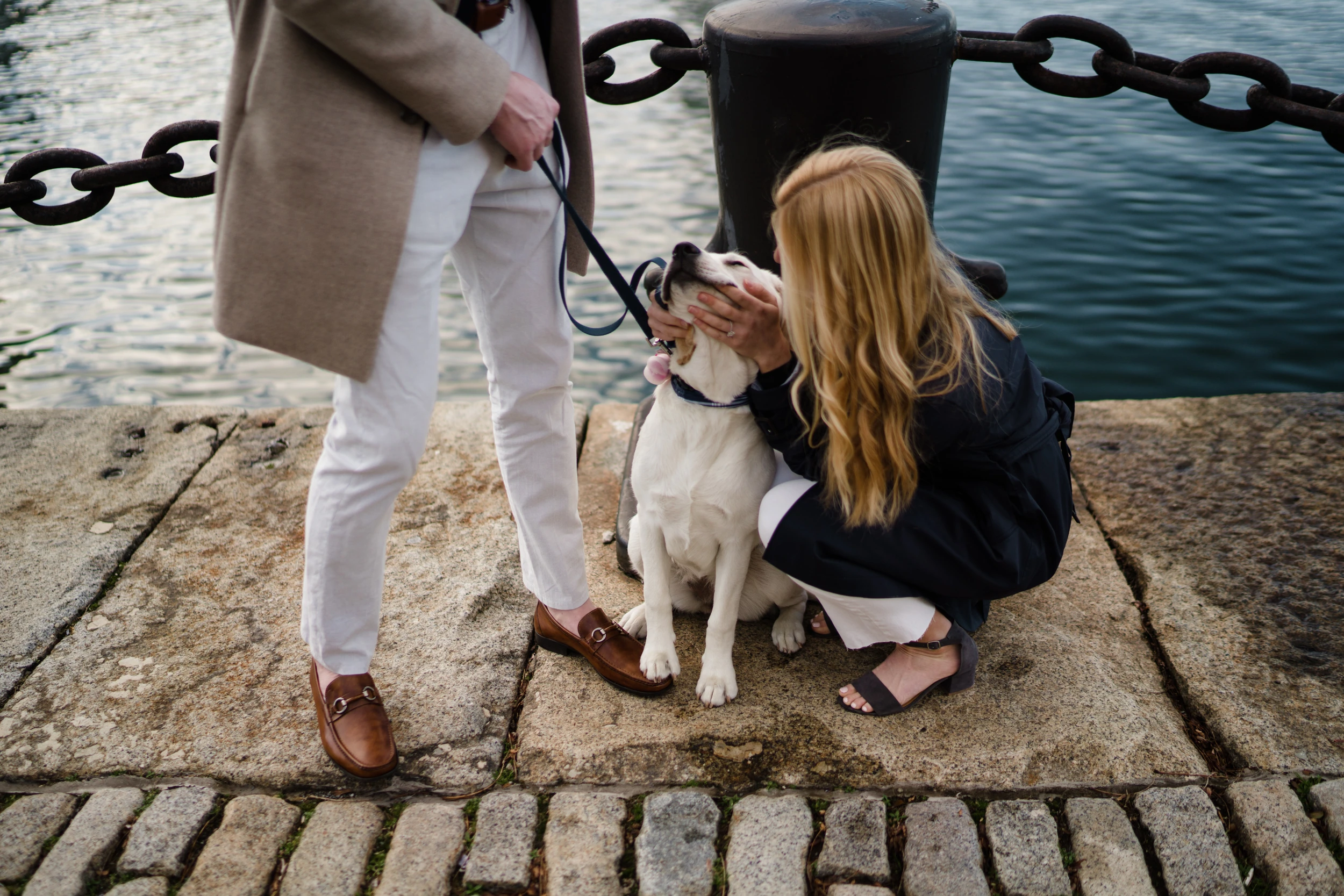 Woman crouches to hug a white dog on a leash at a stone harbour edge, man standing cropped at the waist, chain railing and water behind