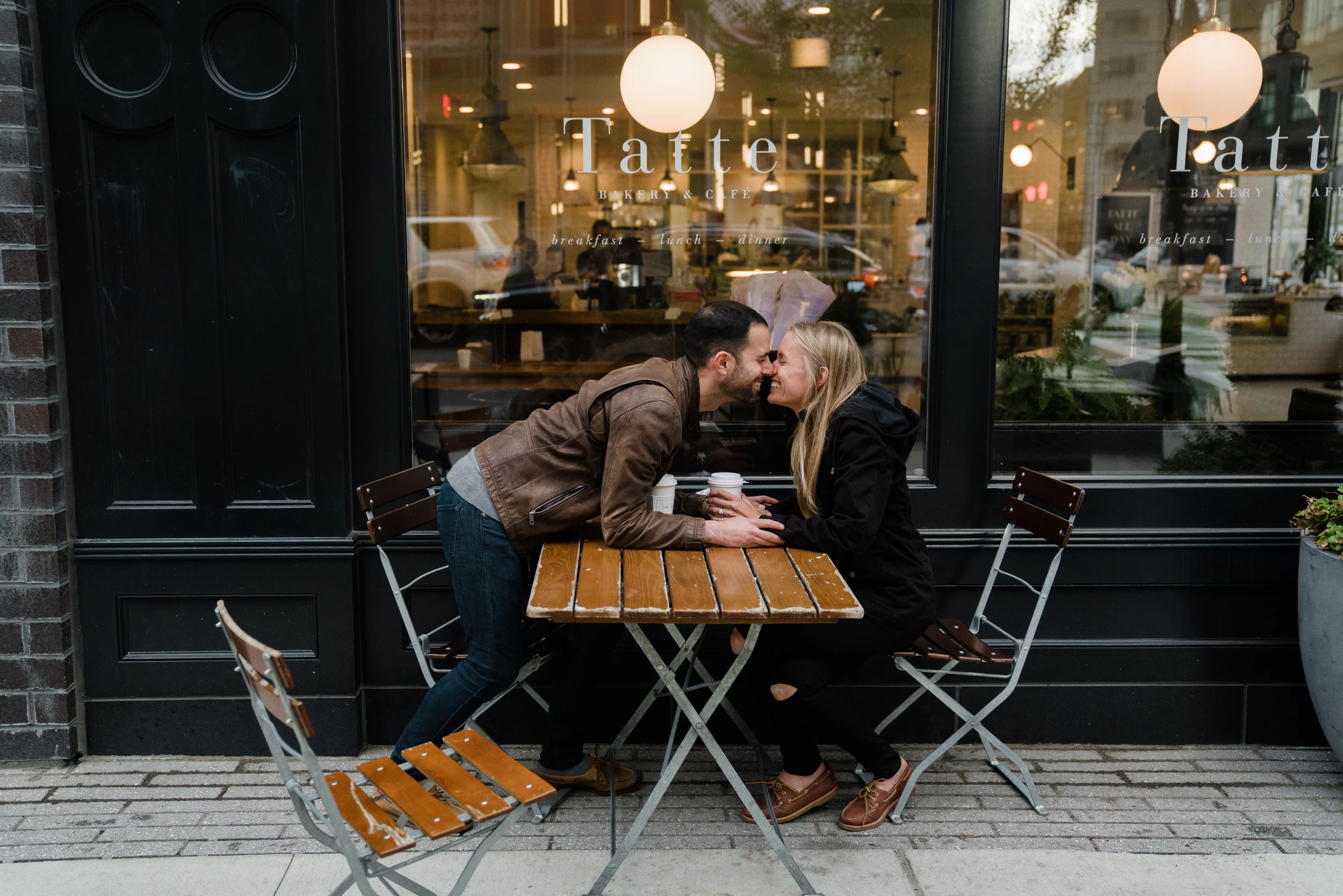 Couple leaning across a small café table to touch noses outside a bakery window, globe lamps above