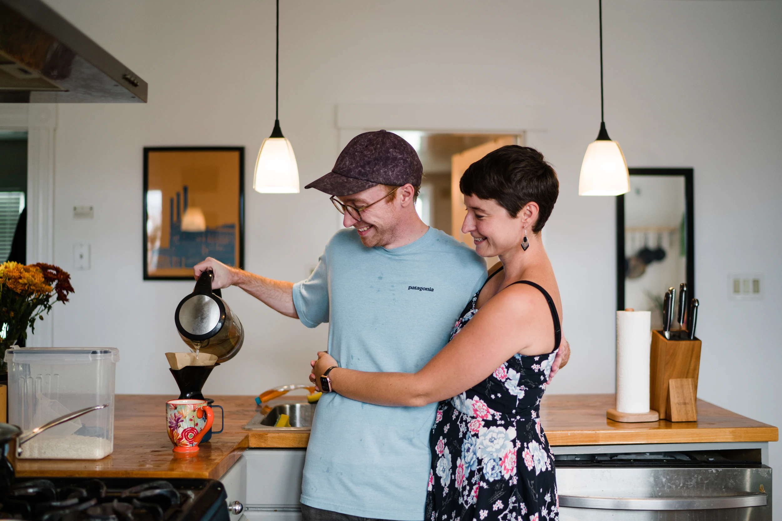 Man pours a kettle over a coffee filter while the woman hugs him from the side in a white kitchen
