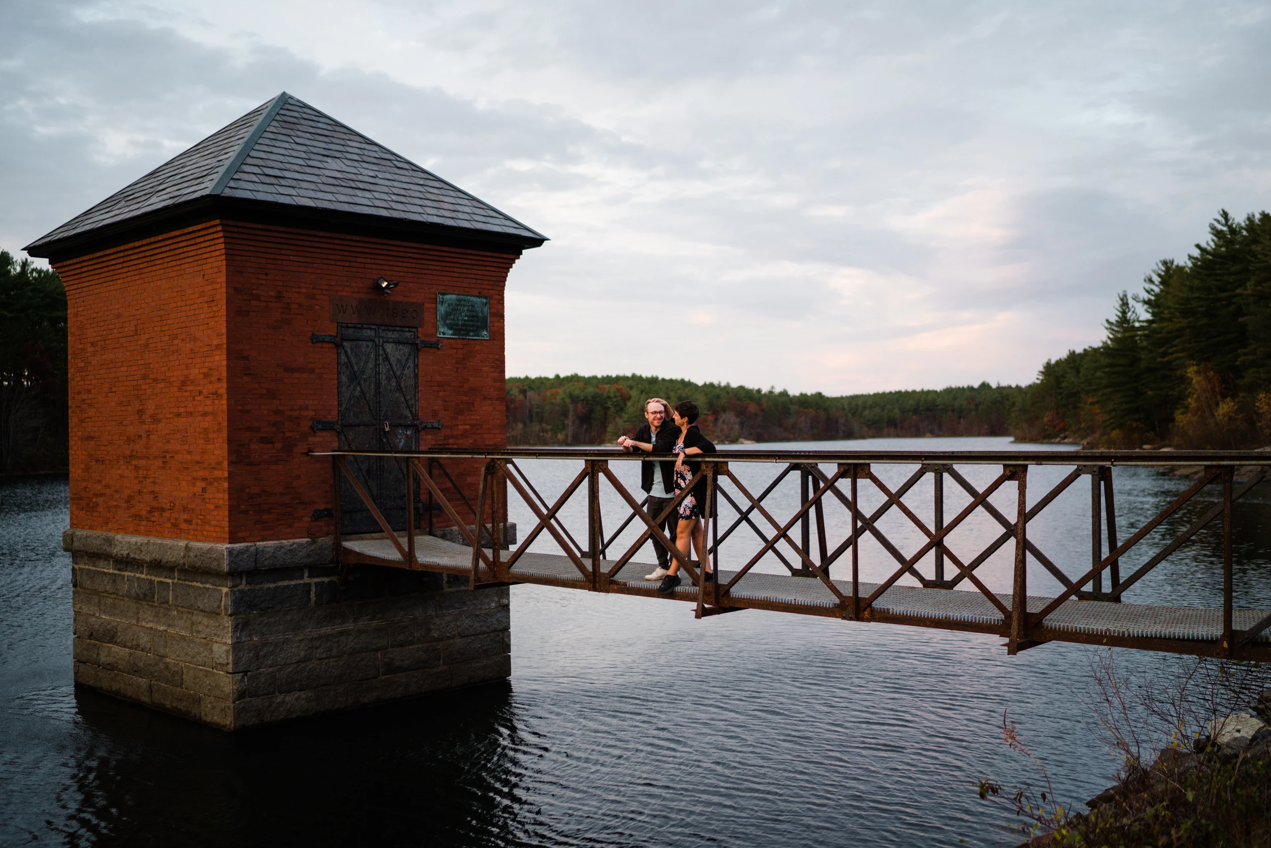 Couple standing on a rusted footbridge to a brick pump house on a reservoir, wide overcast sky