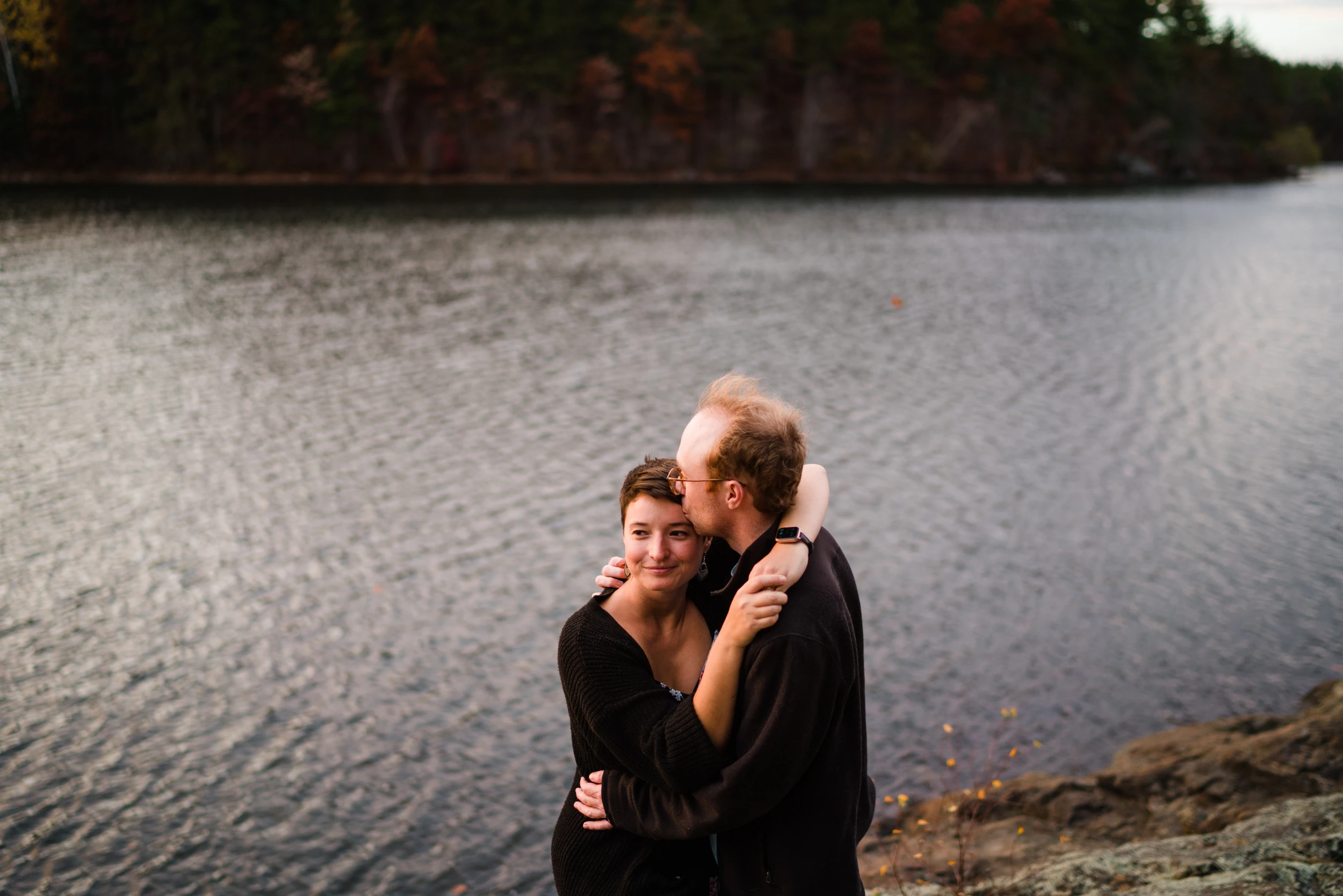 Man kisses the woman's head as they embrace on a rocky shore, rippled lake water filling the frame behind