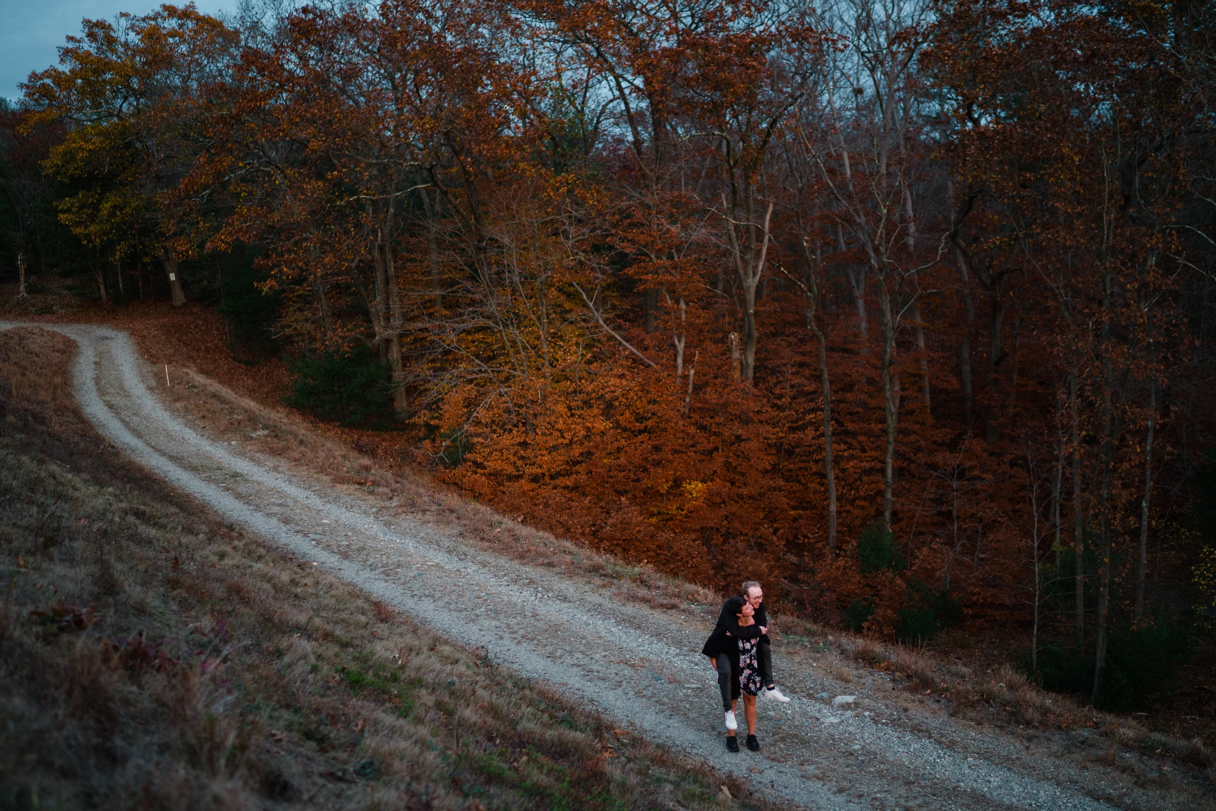 High view of a couple, one piggybacking the other, on a winding gravel road beside autumn woods at dusk