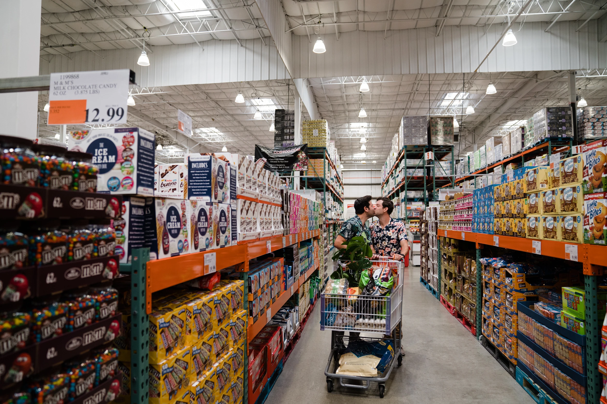Two men kiss behind a loaded shopping cart in a warehouse-store aisle stacked with candy and snack boxes