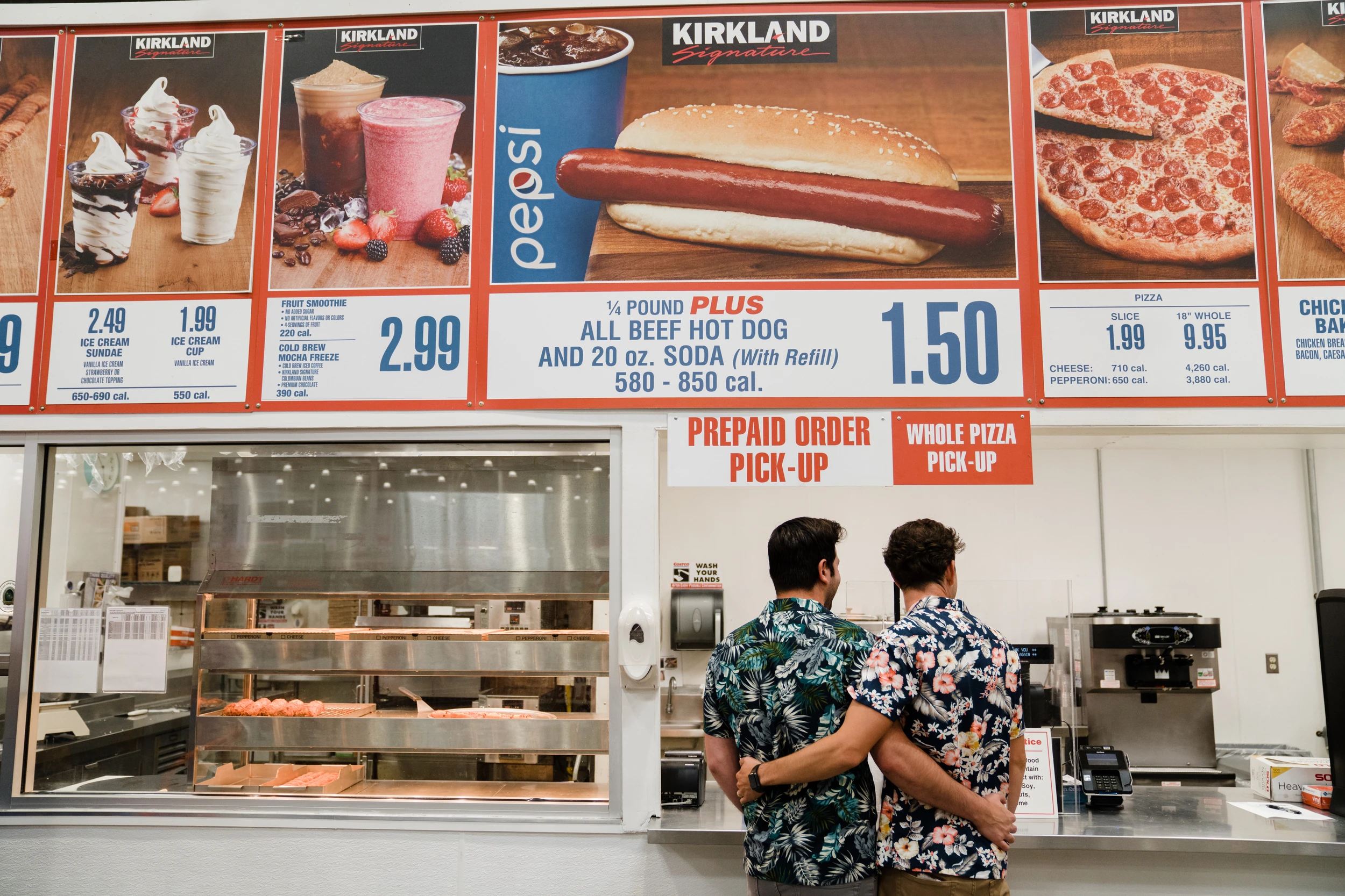 Two men in floral shirts, arms around each other, facing a food-court counter under a large menu board