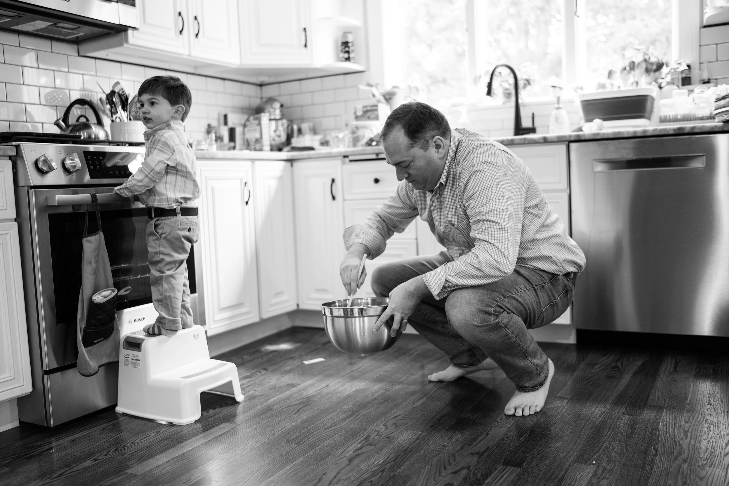 Father squats on the kitchen floor stirring a mixing bowl while a toddler stands on a step stool at the oven