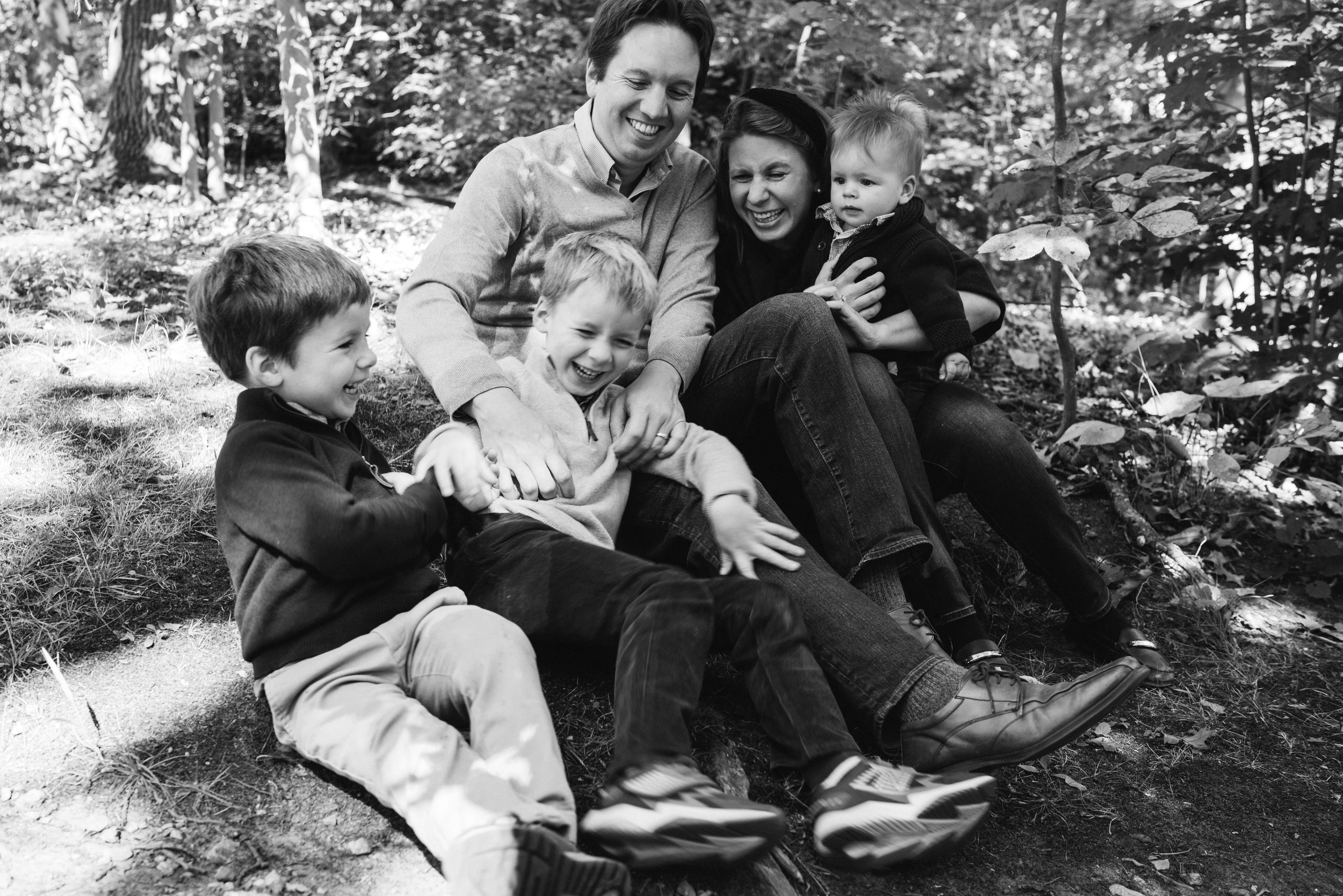 Parents and three young boys laughing in a pile on a wooded hillside in dappled light