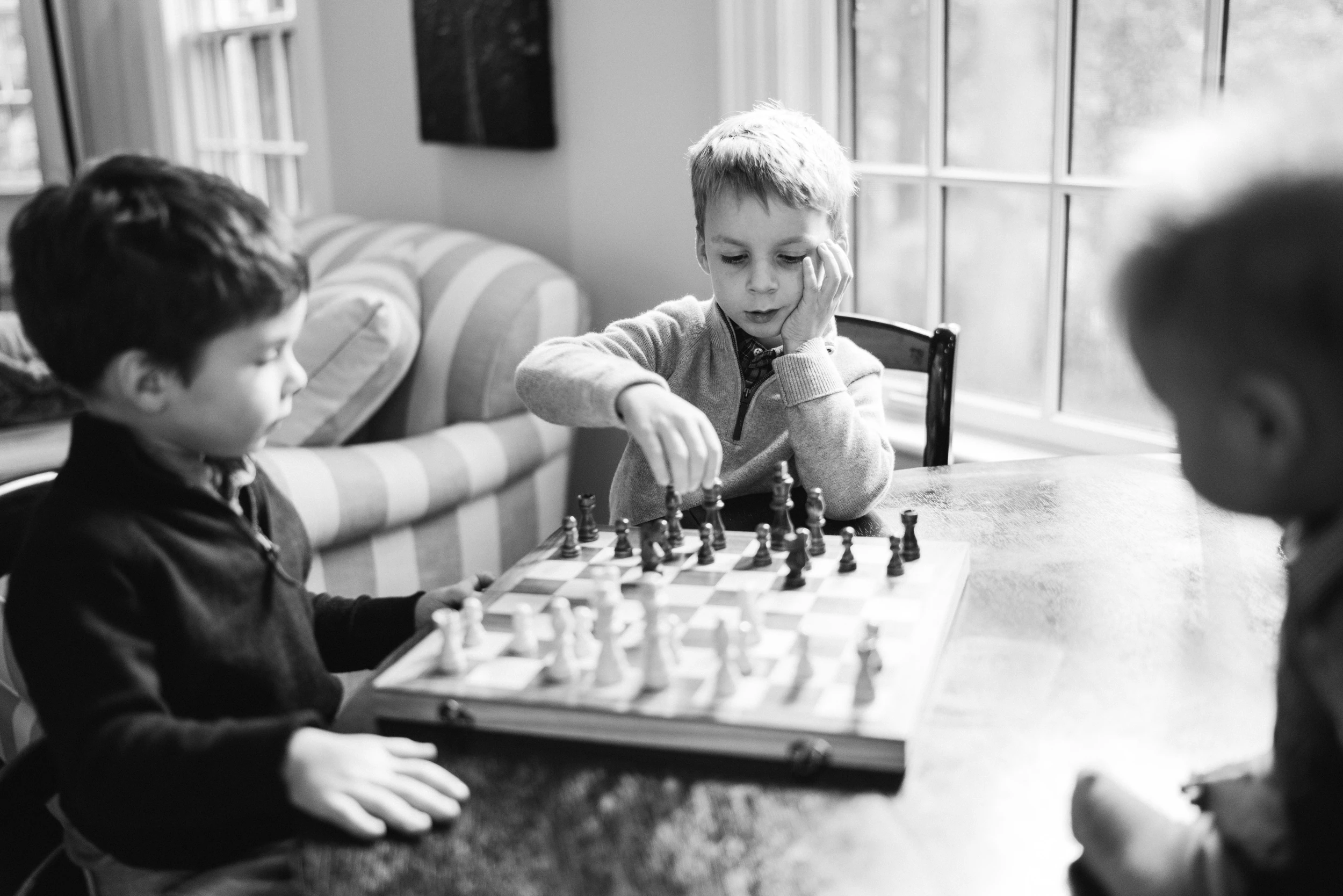 Two boys playing chess at a table by a window, a third child blurred in the foreground