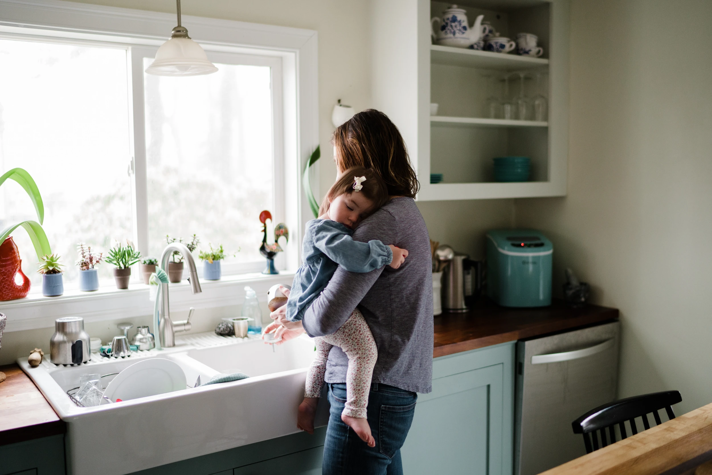 Mother holds a sleeping toddler against her shoulder at a kitchen sink beside a bright window