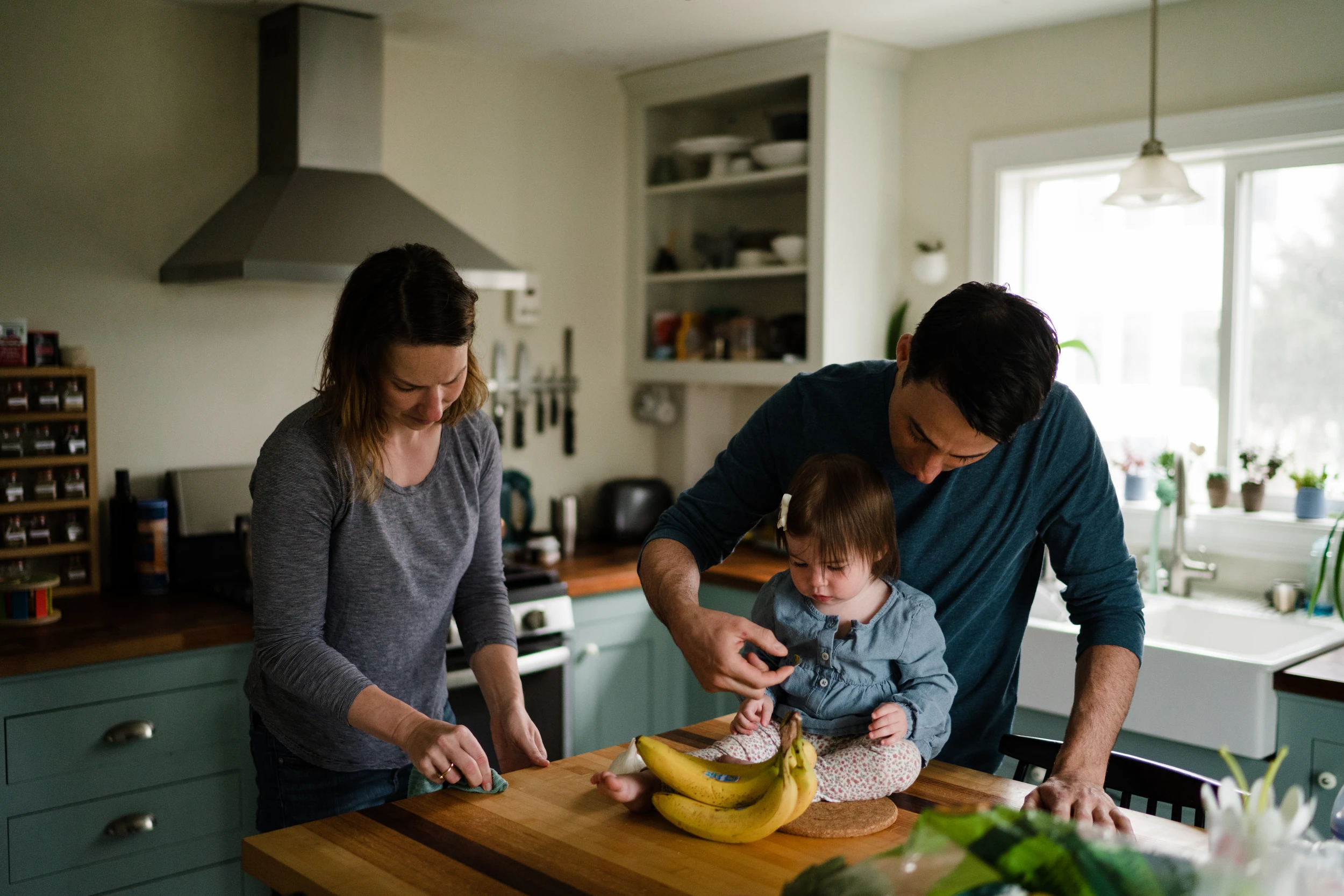 Parents at a butcher-block kitchen island with a toddler sitting on the counter beside a bunch of bananas