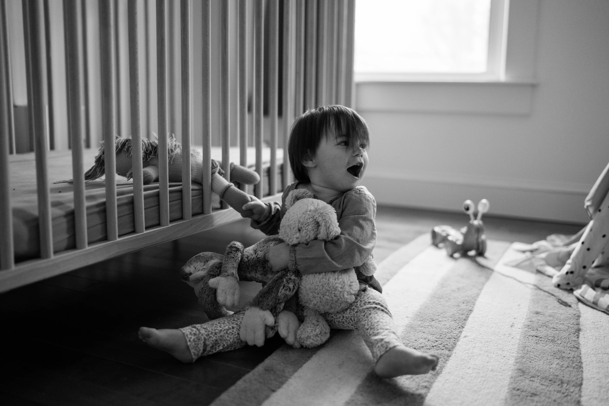 Toddler sitting on a striped rug beside a crib, hugging stuffed animals, mouth open mid-shout