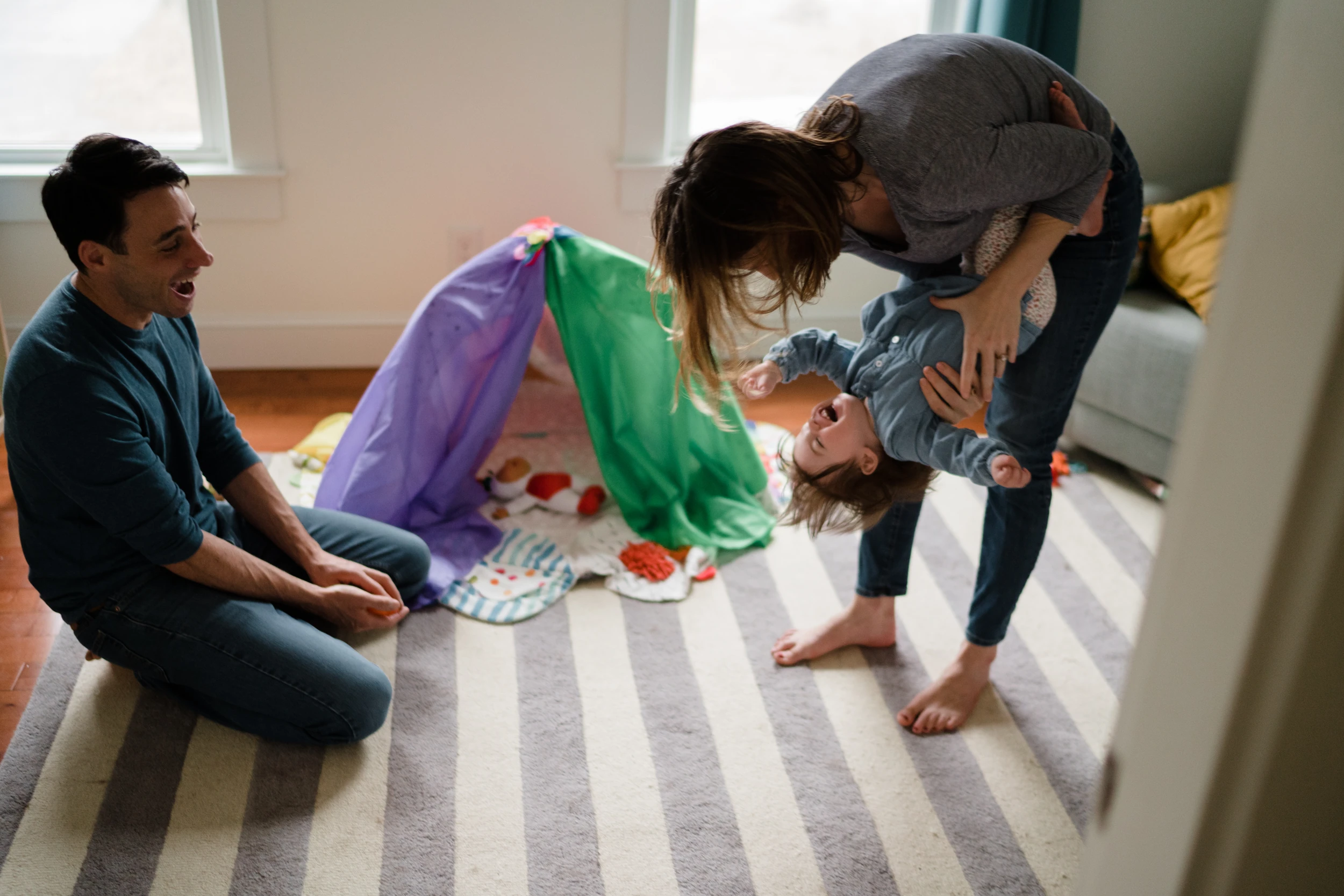 Mother swings a laughing toddler upside down by a play tent while the father kneels and laughs on a striped rug