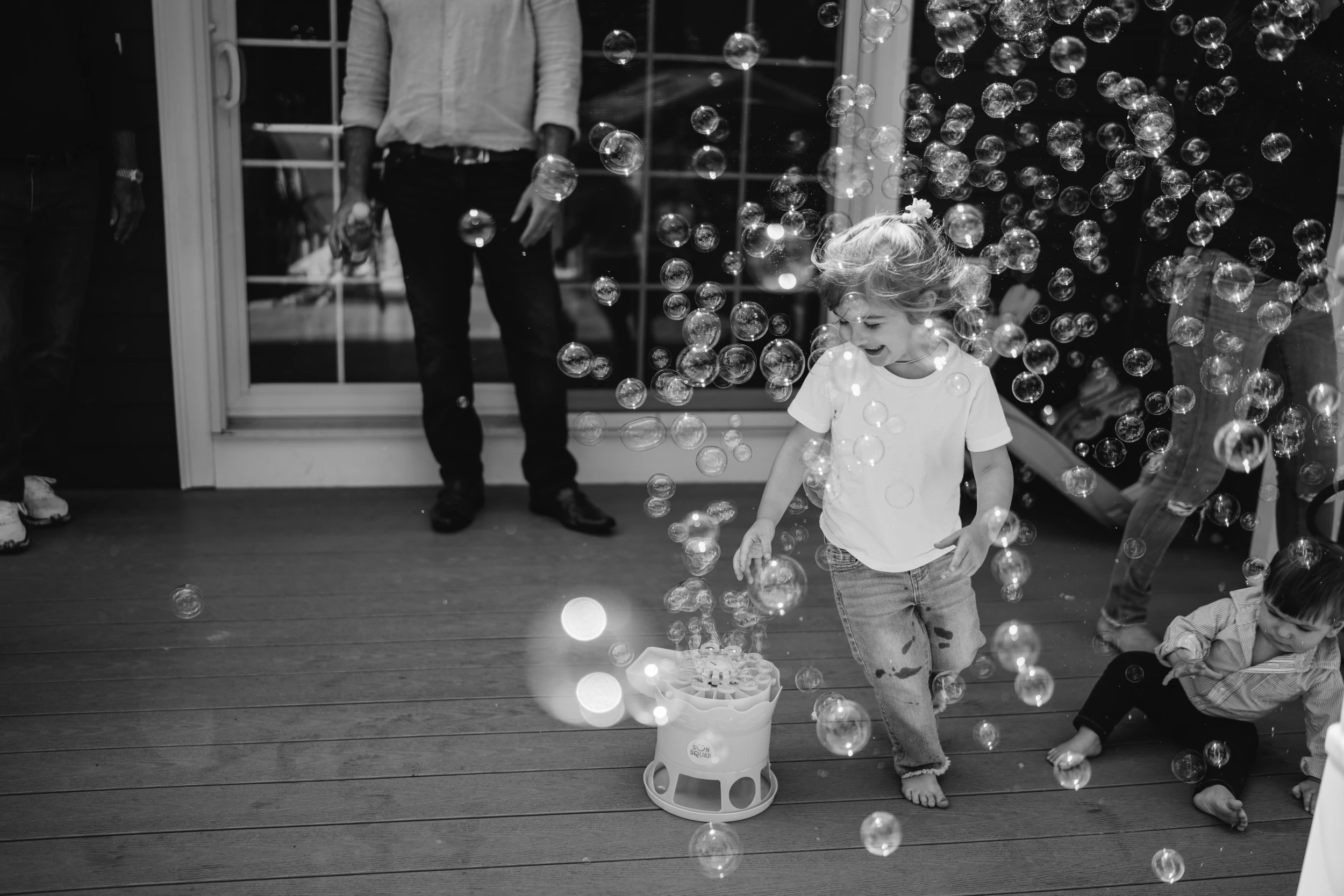 Toddler girl walking through a cloud of soap bubbles on a deck, adults' legs around her