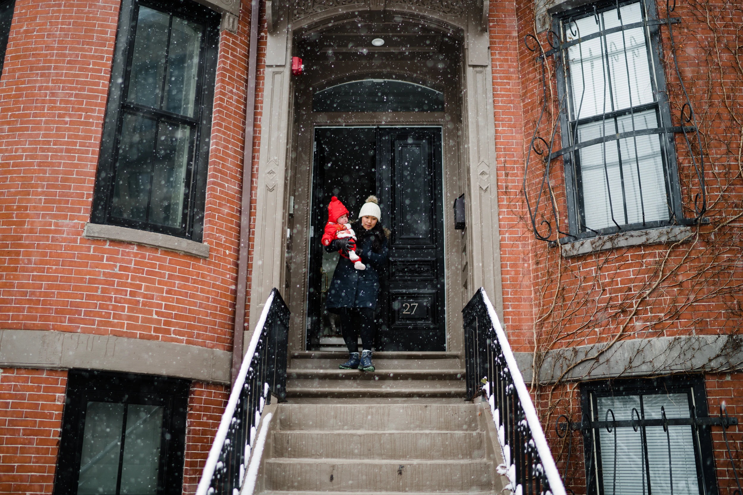 Mother carrying a baby in a red snowsuit out of a black front door onto brownstone steps in falling snow