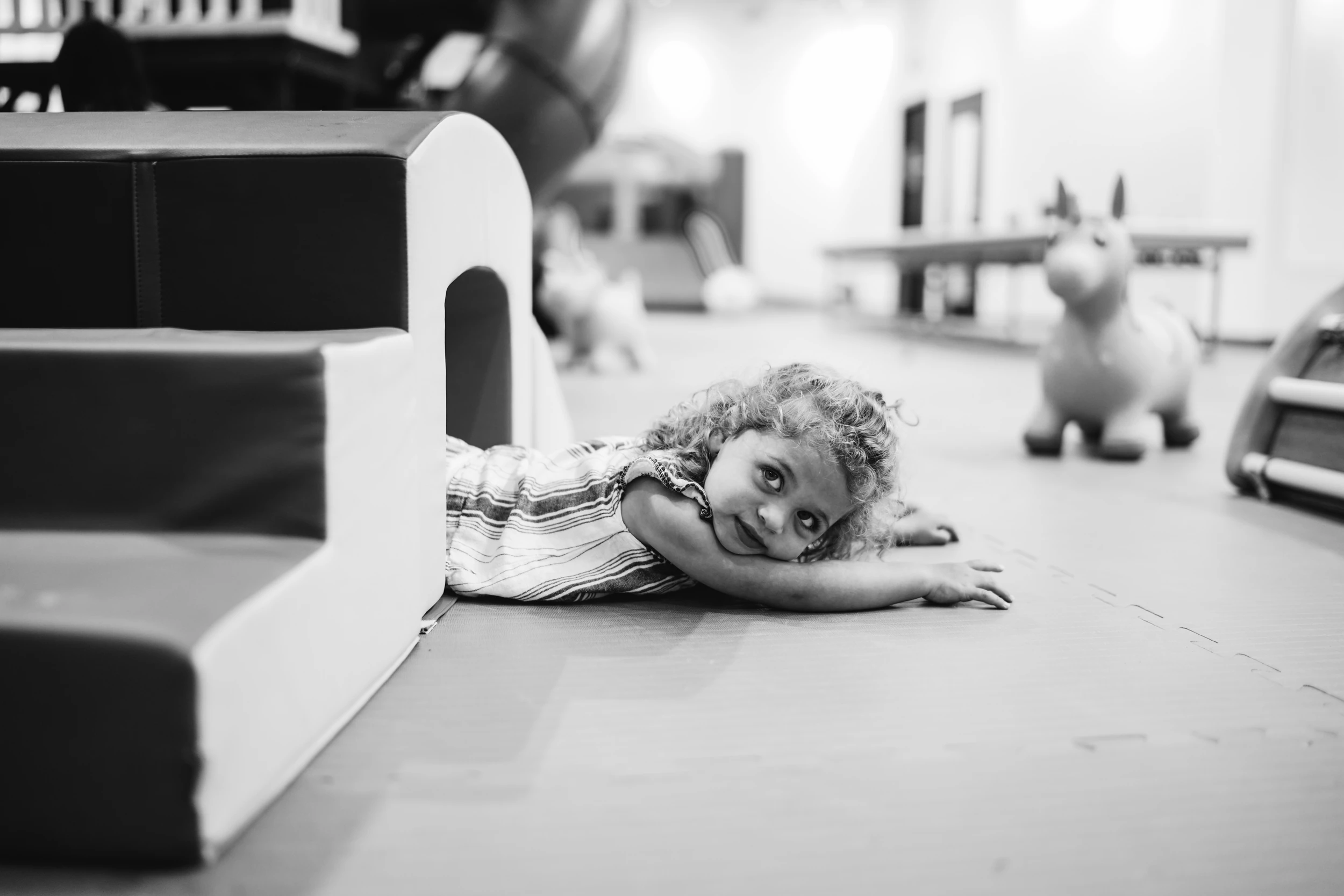 Curly-haired girl lying on a play-gym floor beside foam steps, chin on her arm, looking up at the camera
