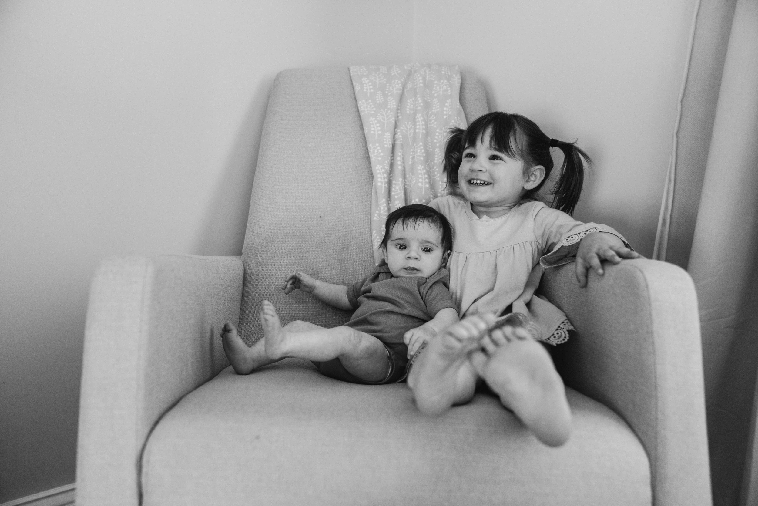 Toddler girl in pigtails and a baby sitting together in a pale armchair against a plain wall