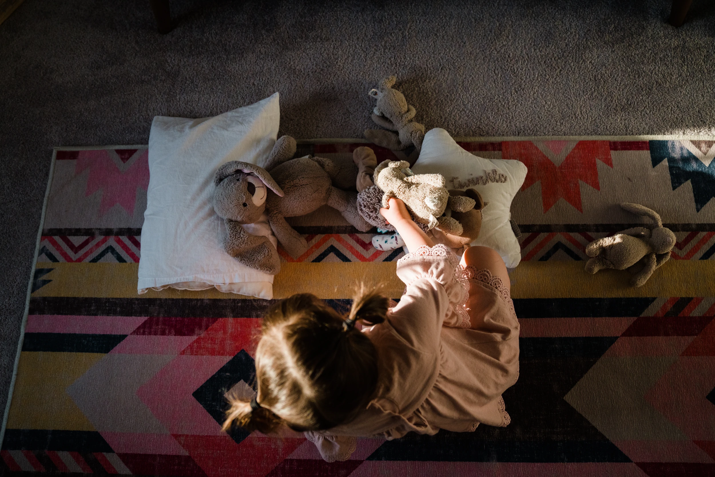 Overhead of a girl in pigtails sitting on a geometric rug arranging stuffed bunnies and pillows