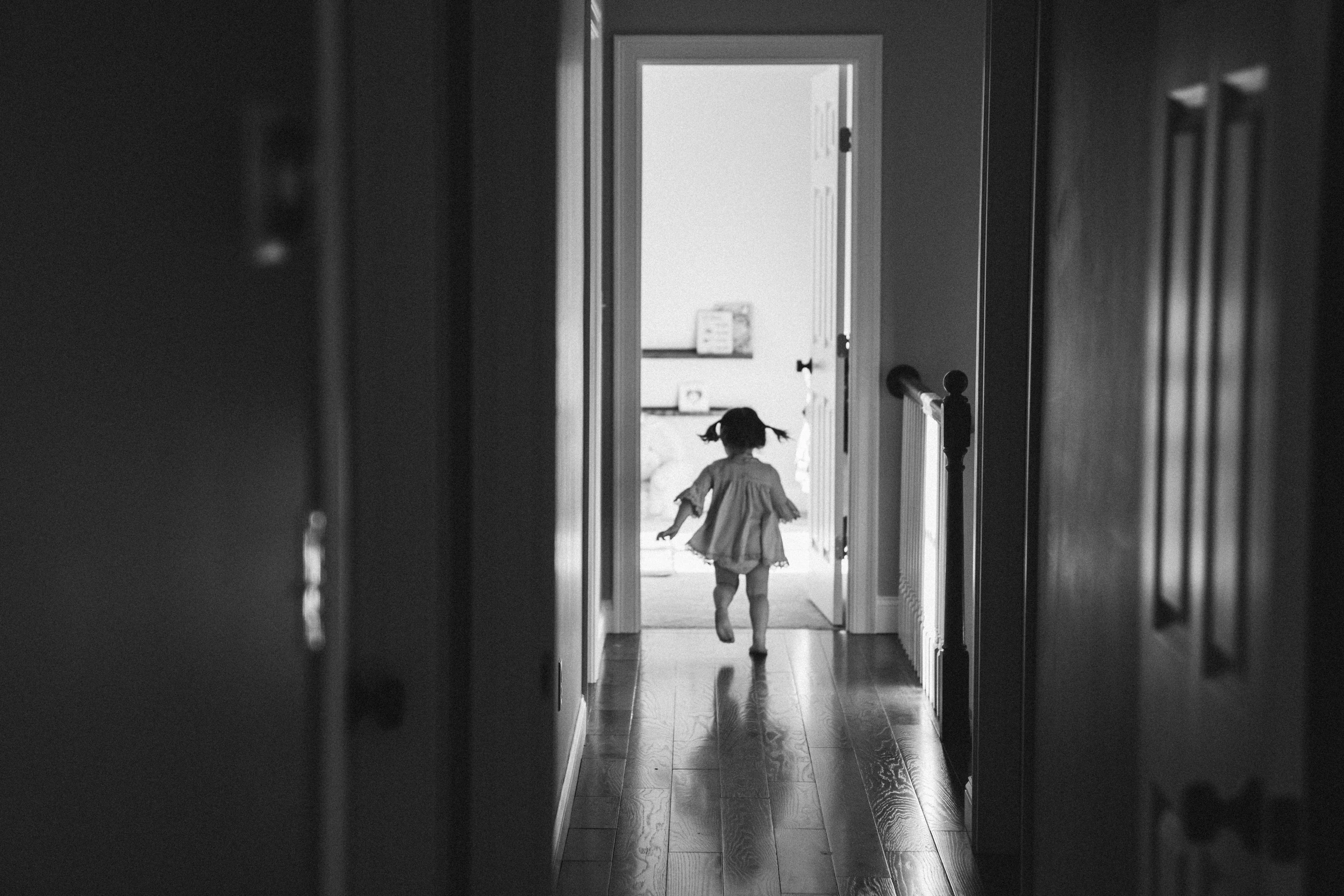 Toddler girl in pigtails running down a dark hallway toward a bright open doorway