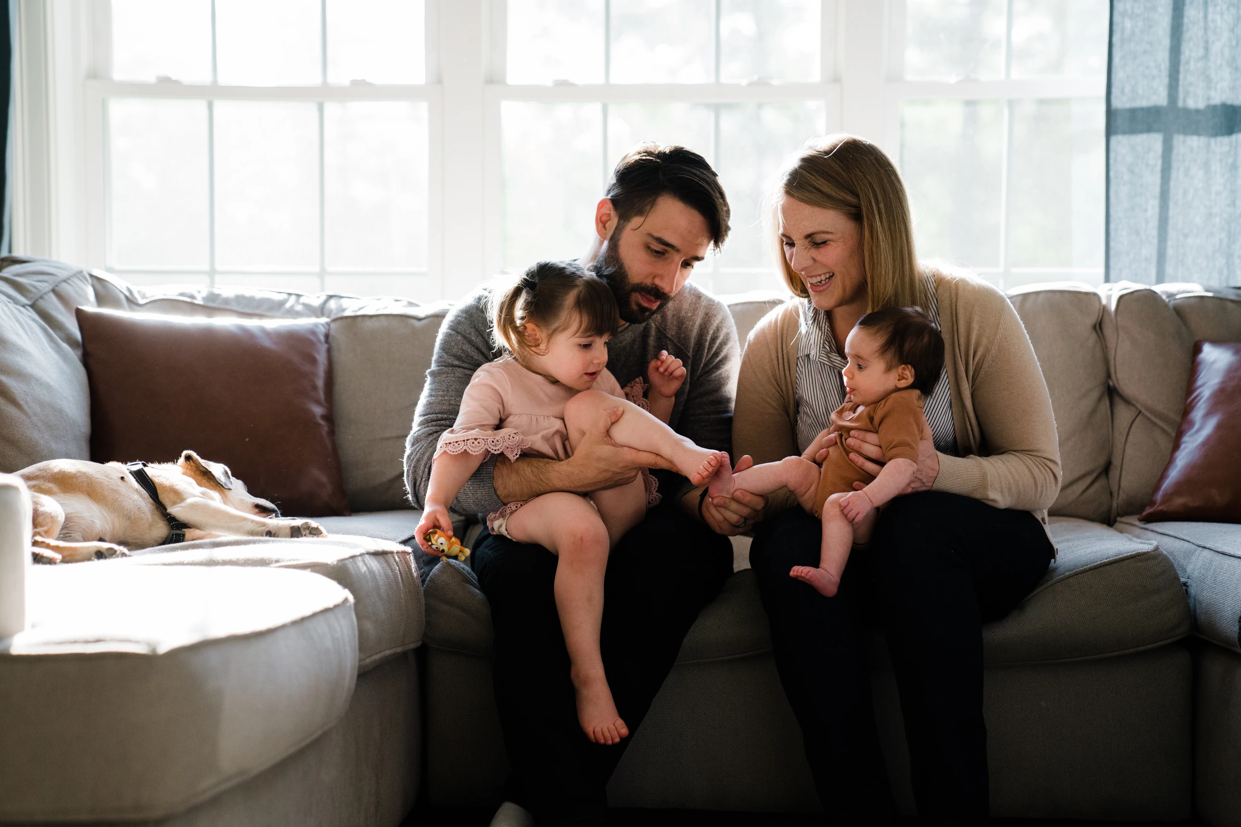 Parents on a sofa holding a toddler and a baby, a dog asleep on the cushion beside them, bright windows behind