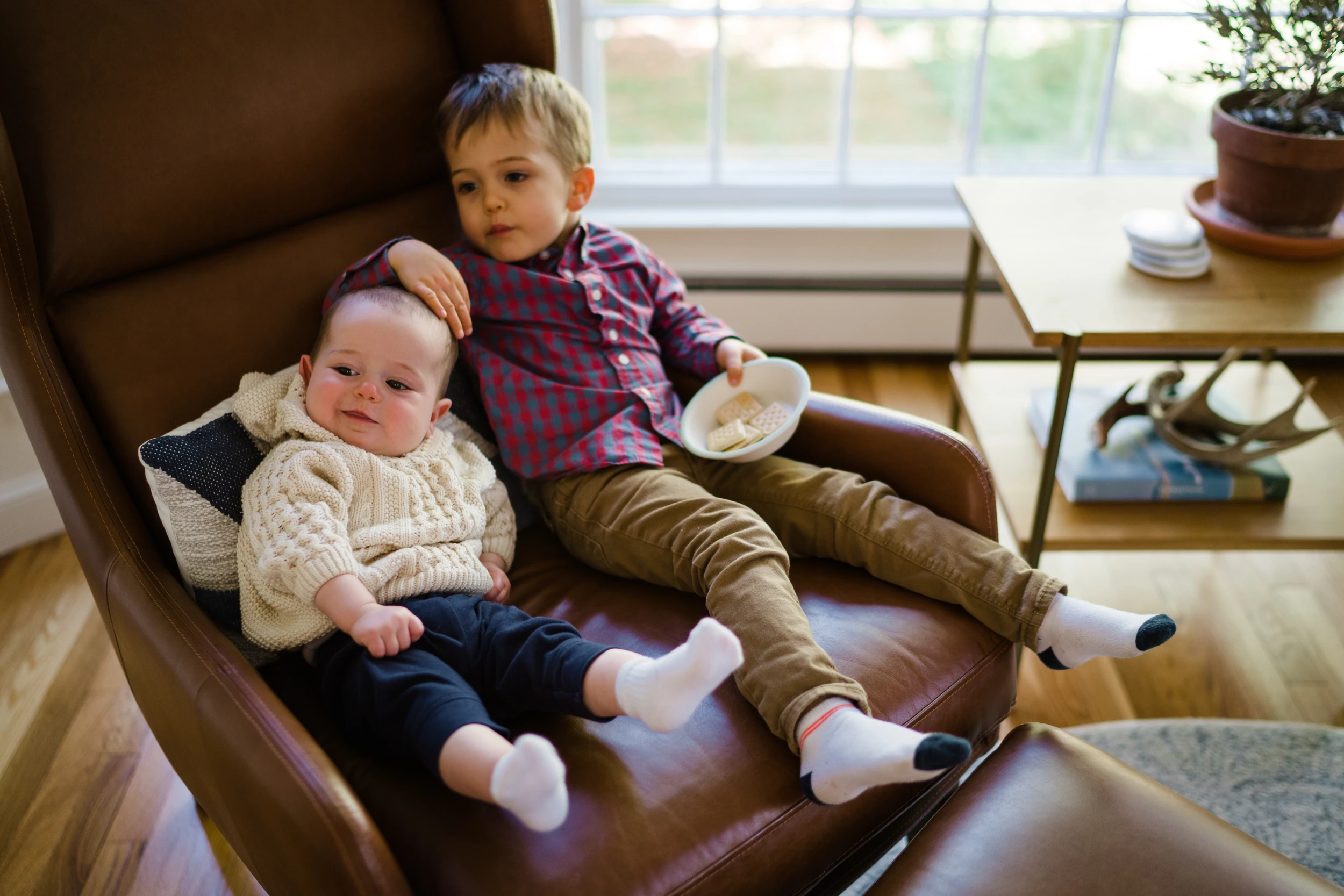 Older boy lying in a leather armchair with his arm around a baby, holding a bowl of snacks