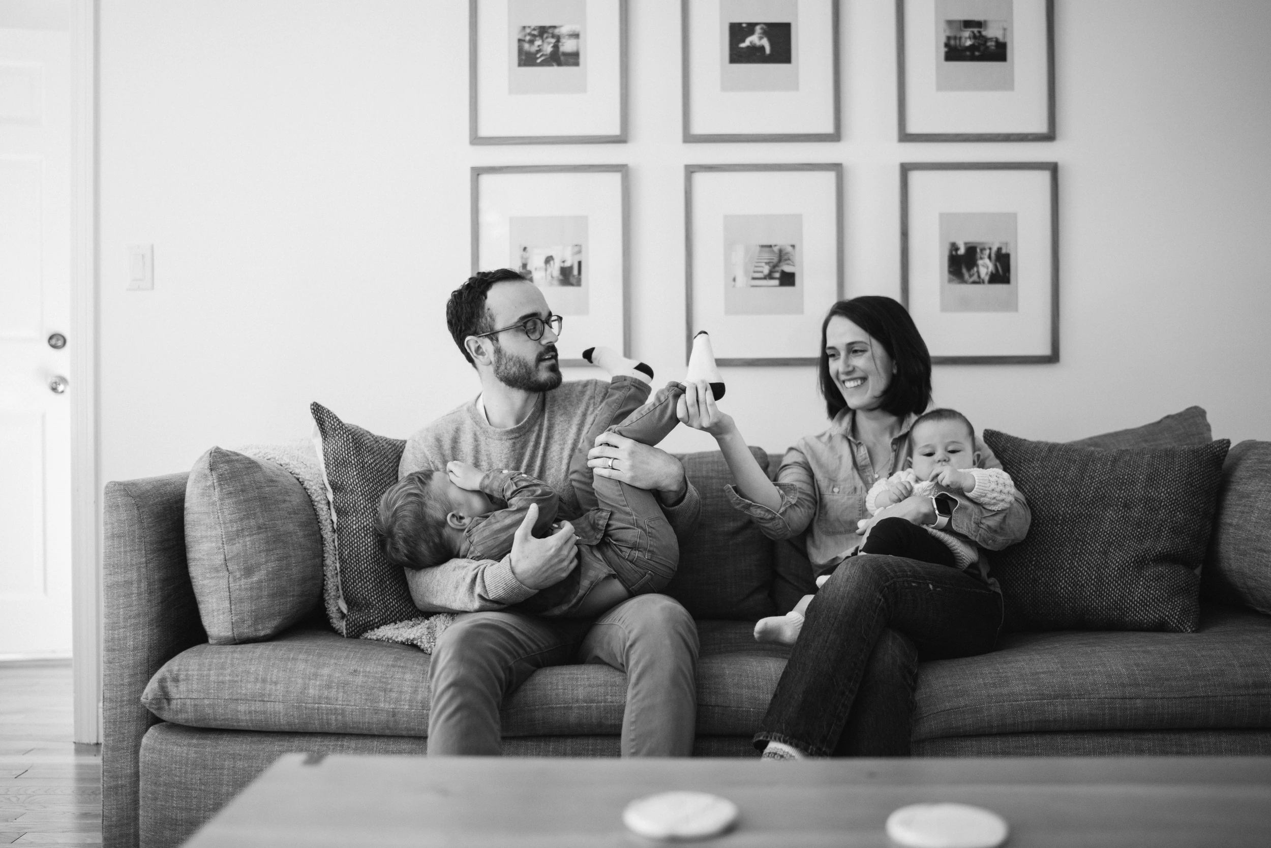 Parents on a sofa each holding a child, a grid of framed photos on the wall above