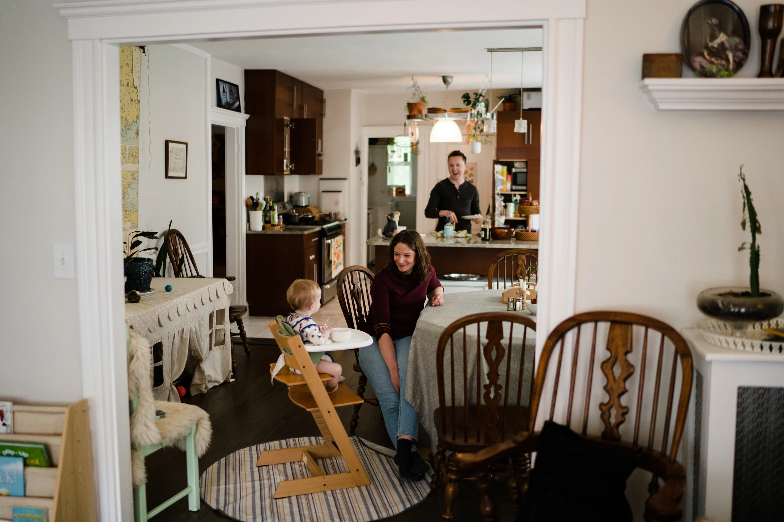 View through a doorway into a kitchen: mother at the table beside a baby in a high chair, father laughing at the counter