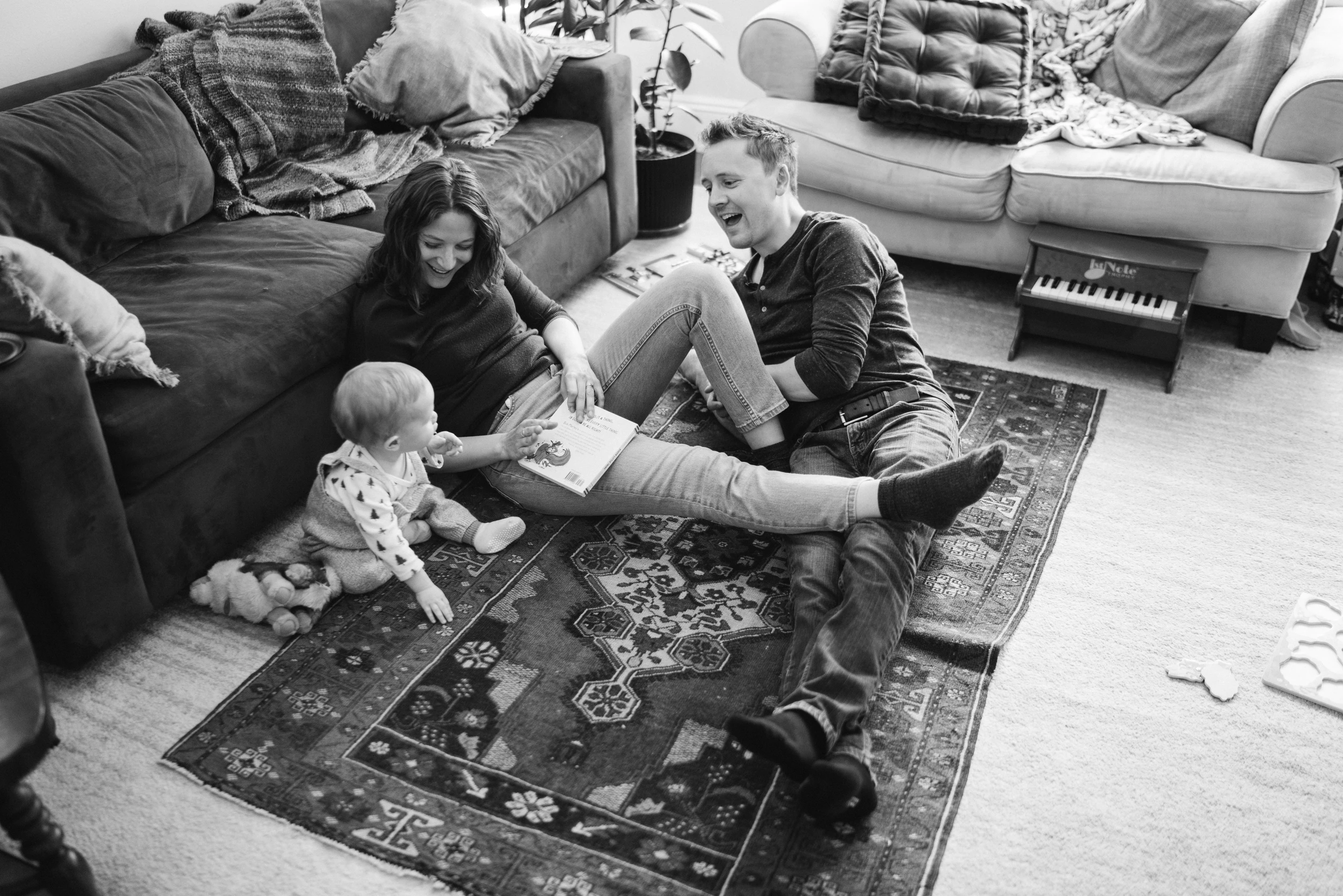 Parents lying back laughing on a patterned rug between sofas as a baby reaches toward a book