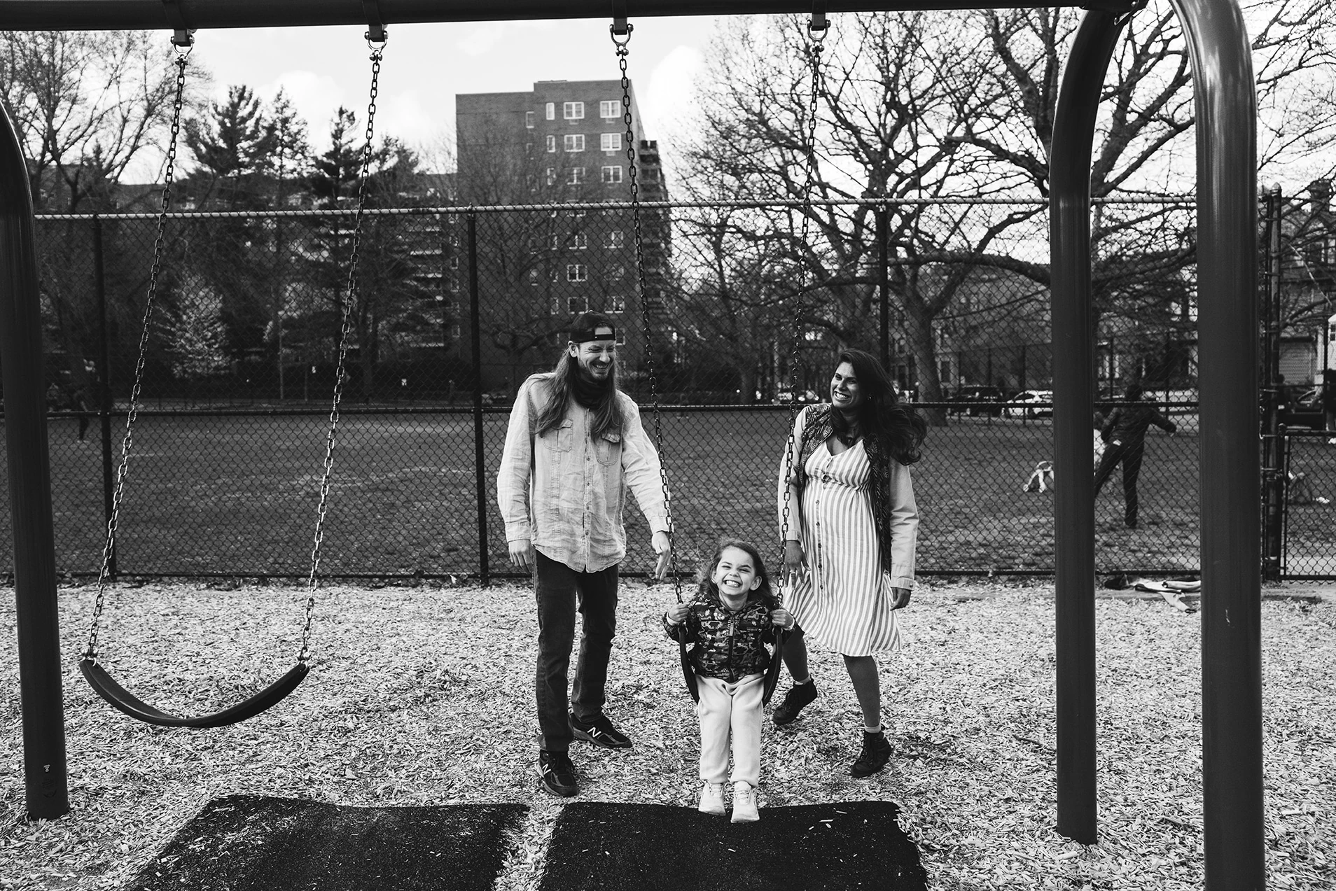 Framed by a swing set, parents walk with a laughing girl between them across a park playground