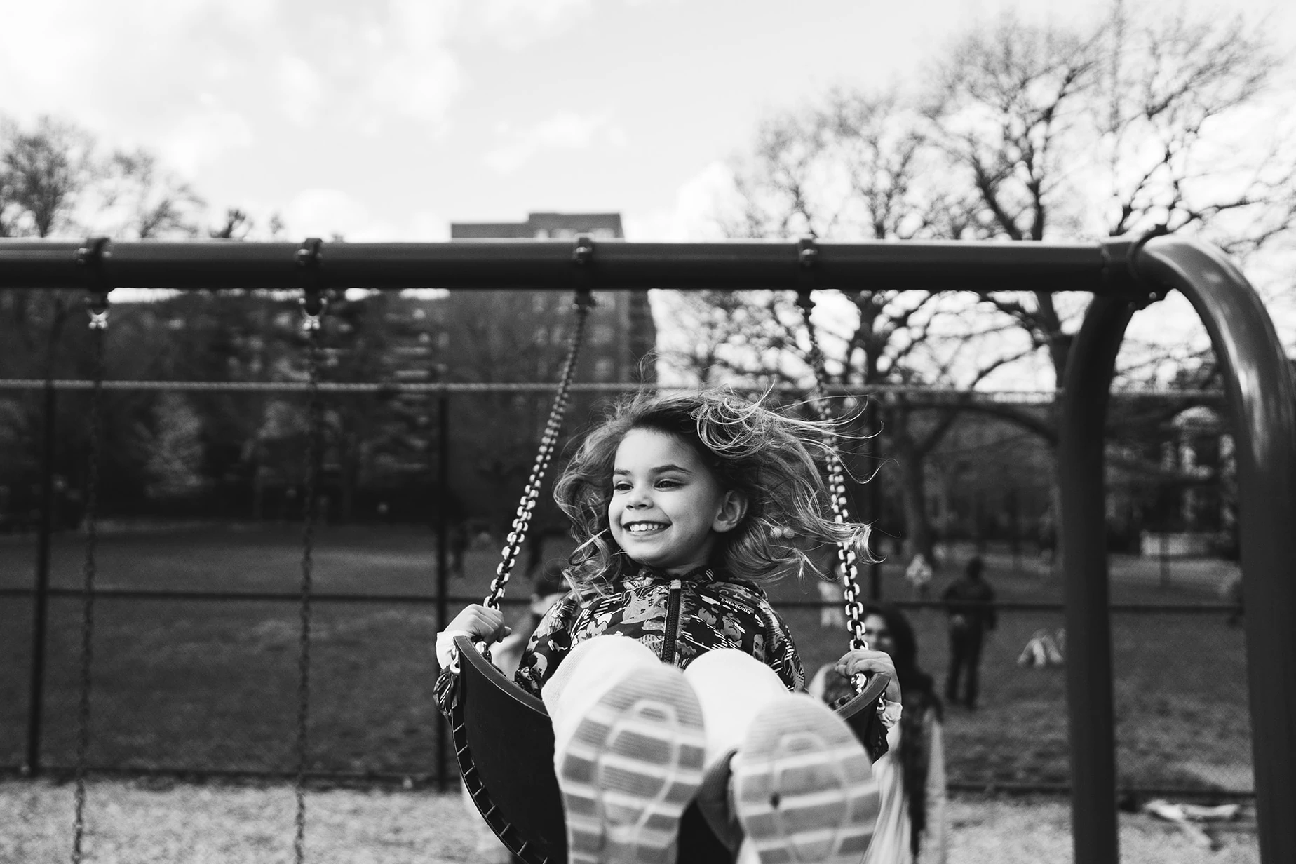 Girl laughing on a playground swing, feet toward the camera, hair flying